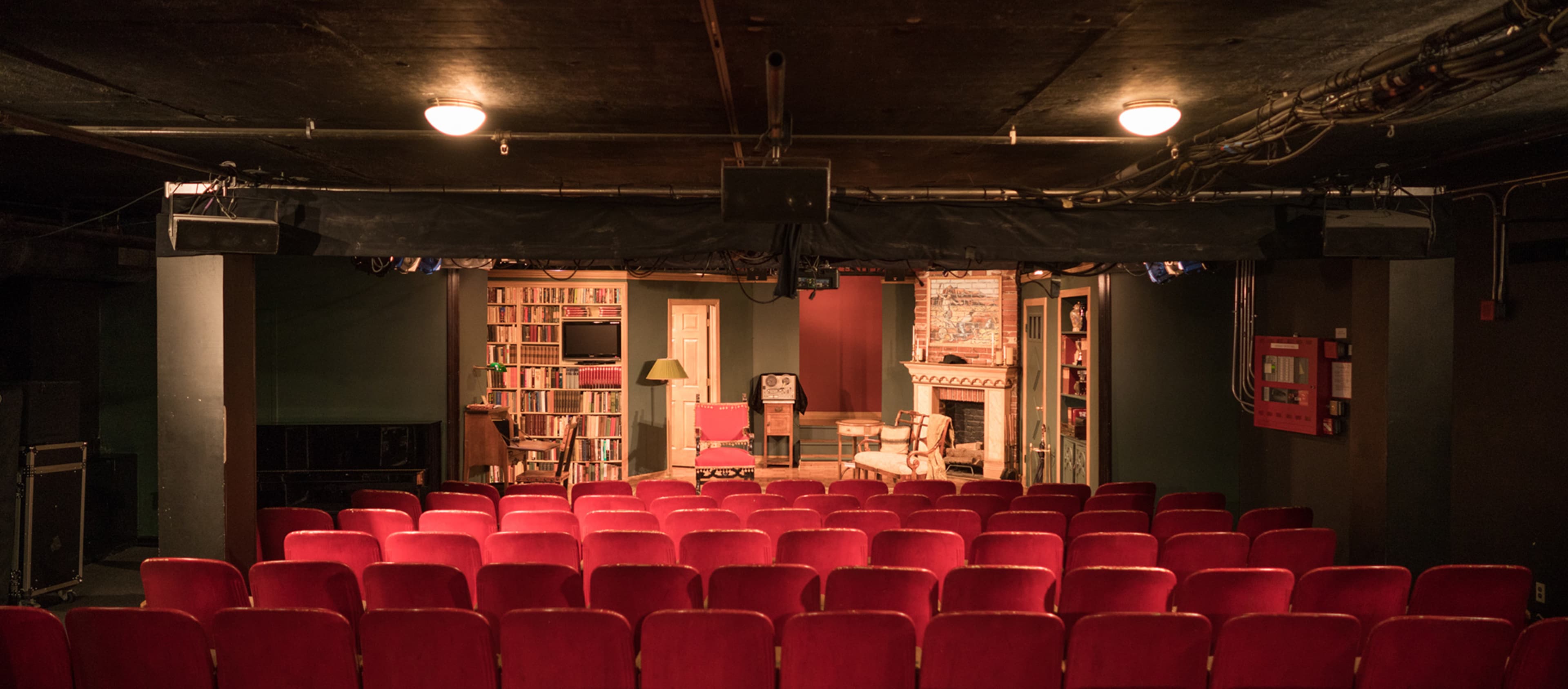 The image shows a small theater with red seats facing a stage that features a living room set, complete with a bookshelf, a lamp, and a fireplace.