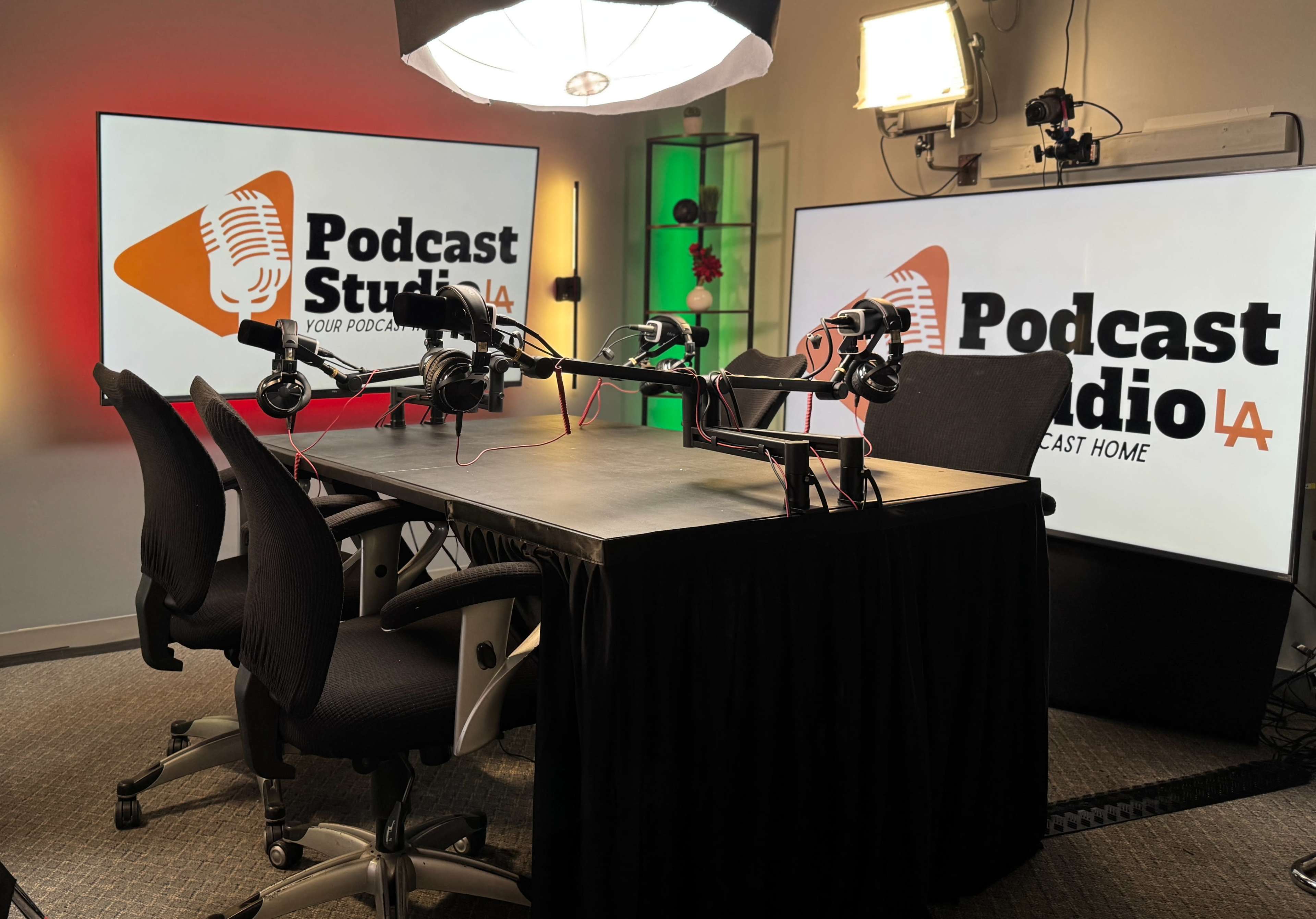 The image shows a podcast studio setup with a table, black chairs, and multiple microphones positioned for recording, accompanied by large screens displaying the text "Podcast Studio LA."