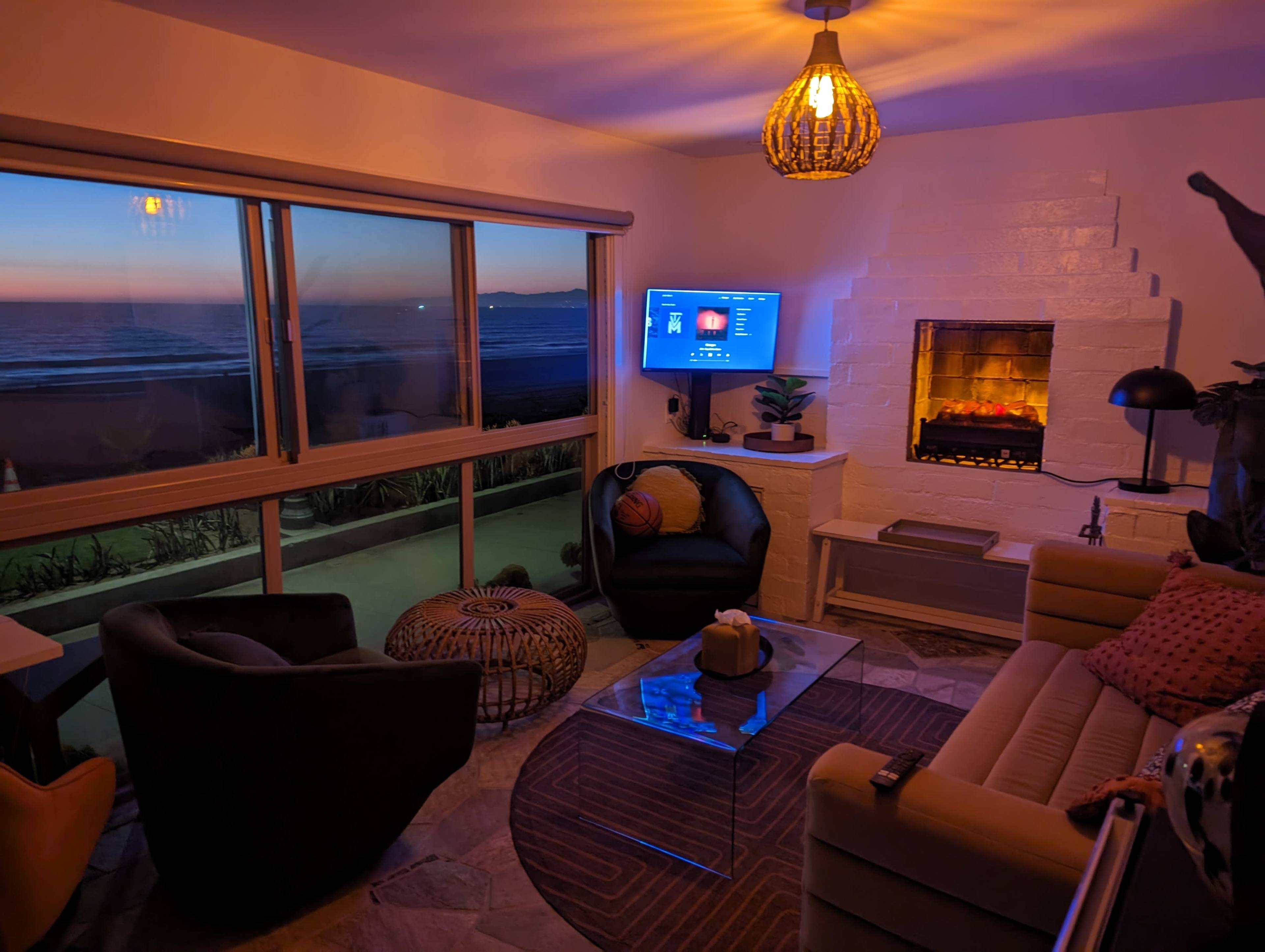 A cozy living room features a modern fireplace, a television, and large windows revealing a sunset by the ocean.