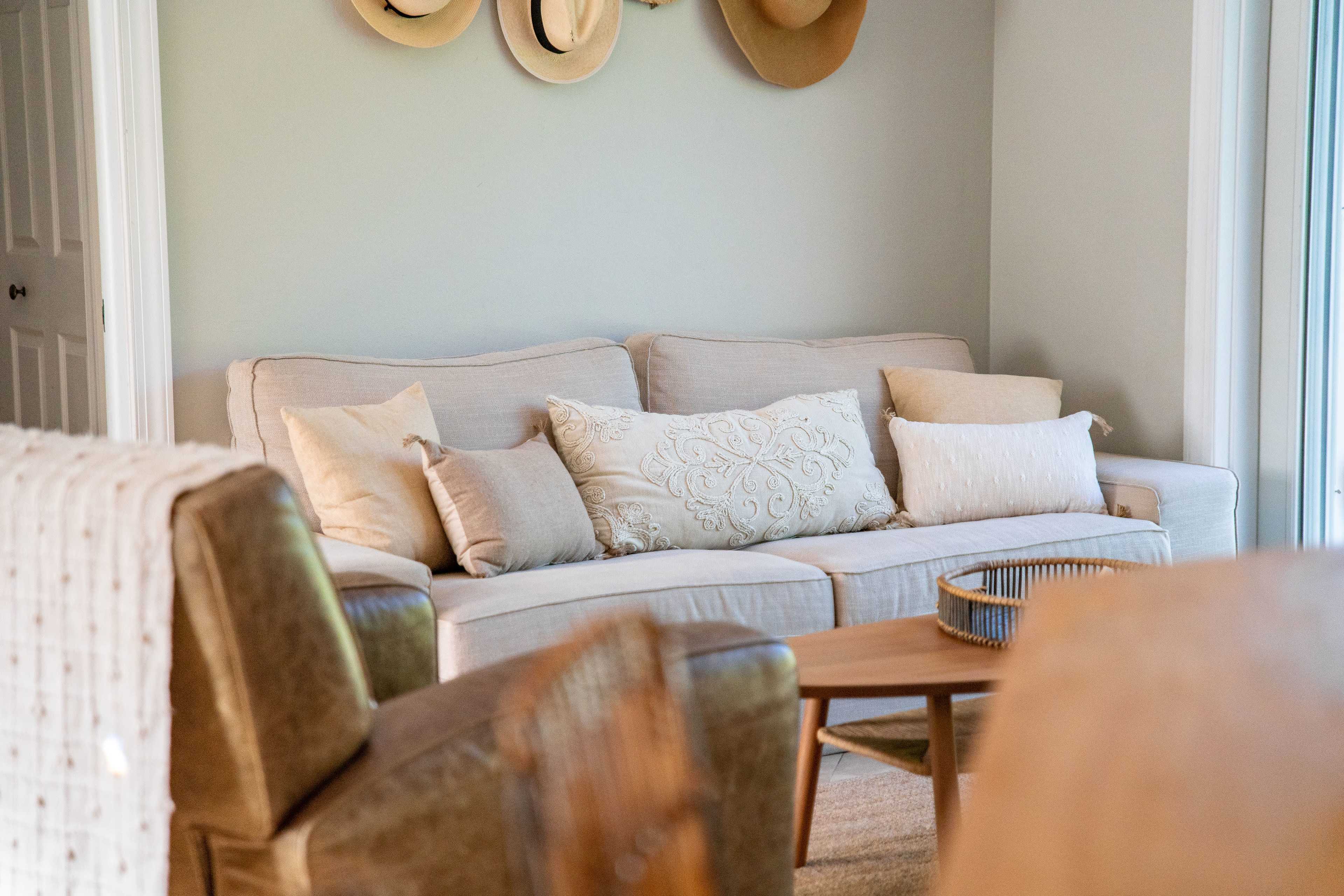 A light-colored sofa with decorative pillows is positioned in a cozy living room, accompanied by a wooden coffee table and a wall adorned with hats.