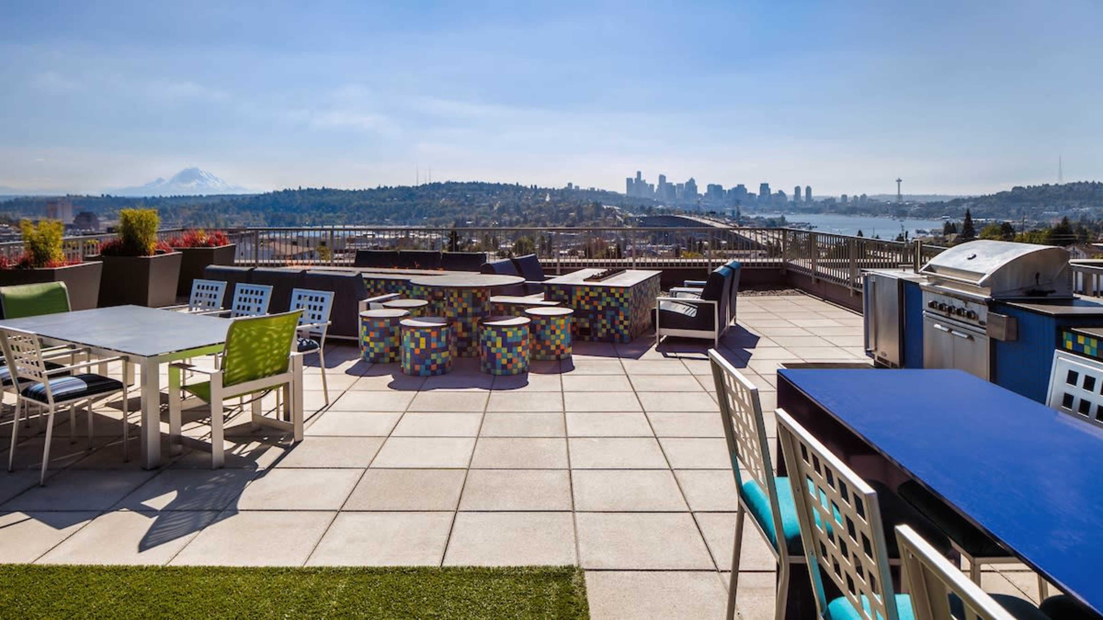 A rooftop terrace features outdoor furniture, a barbecue grill, and a view of a city skyline and mountains in the background.