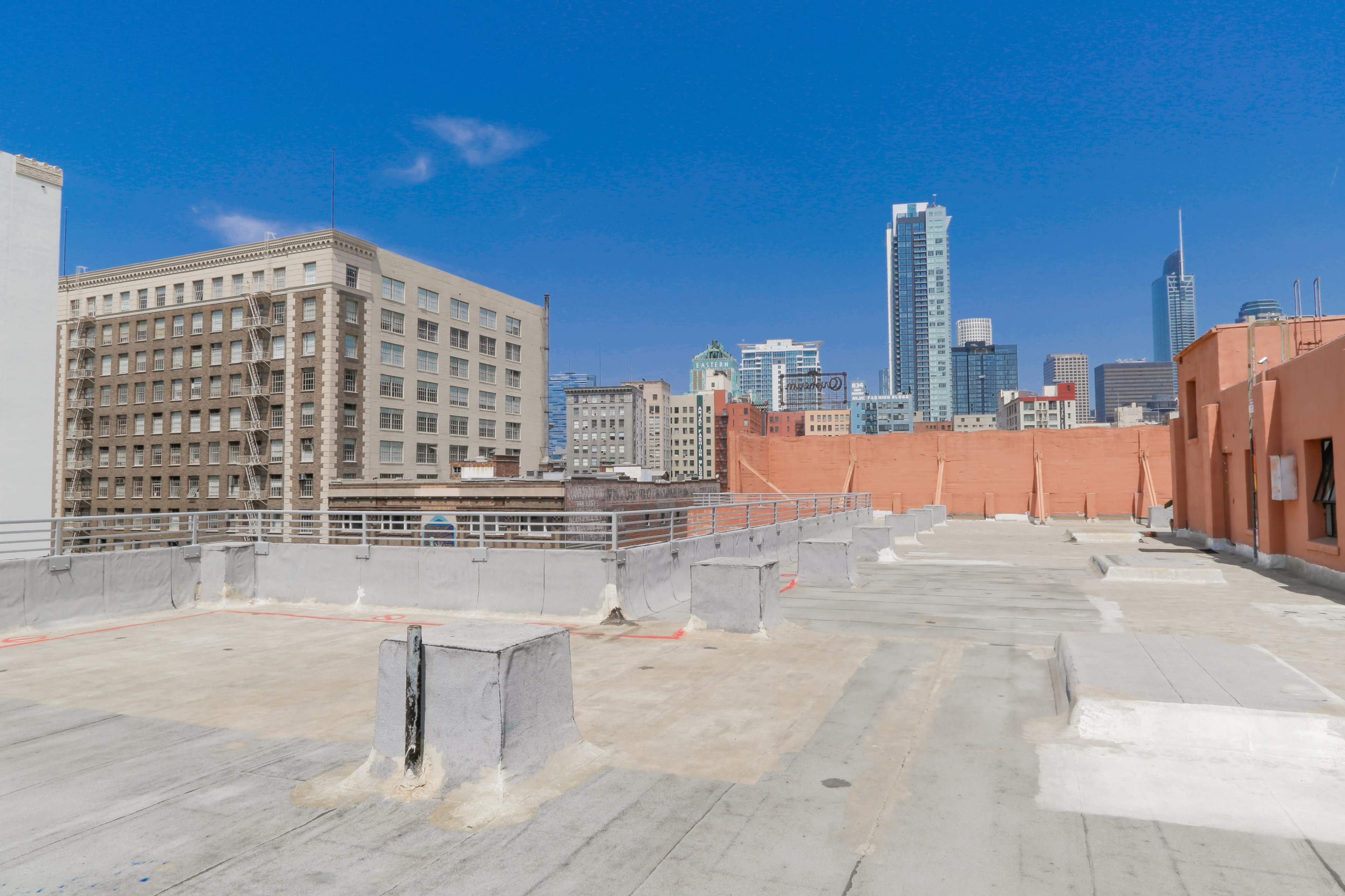 The image shows a rooftop with a flat surface, surrounded by urban buildings and a skyline of skyscrapers against a clear blue sky.