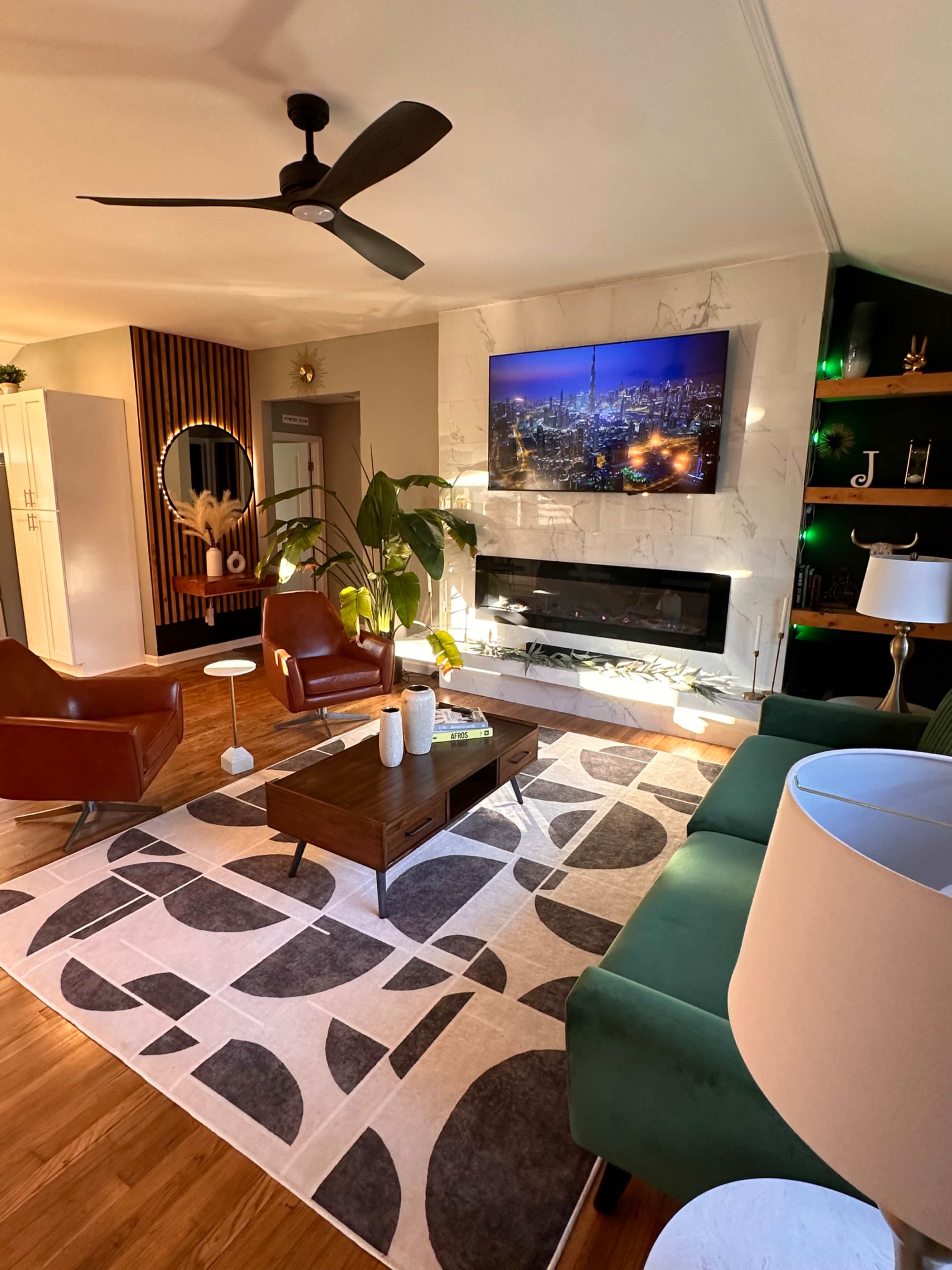 The living room features a modern layout with two brown chairs, a green sofa, a wooden coffee table, and a wall-mounted television above a white fireplace, all arranged on a geometric patterned rug.