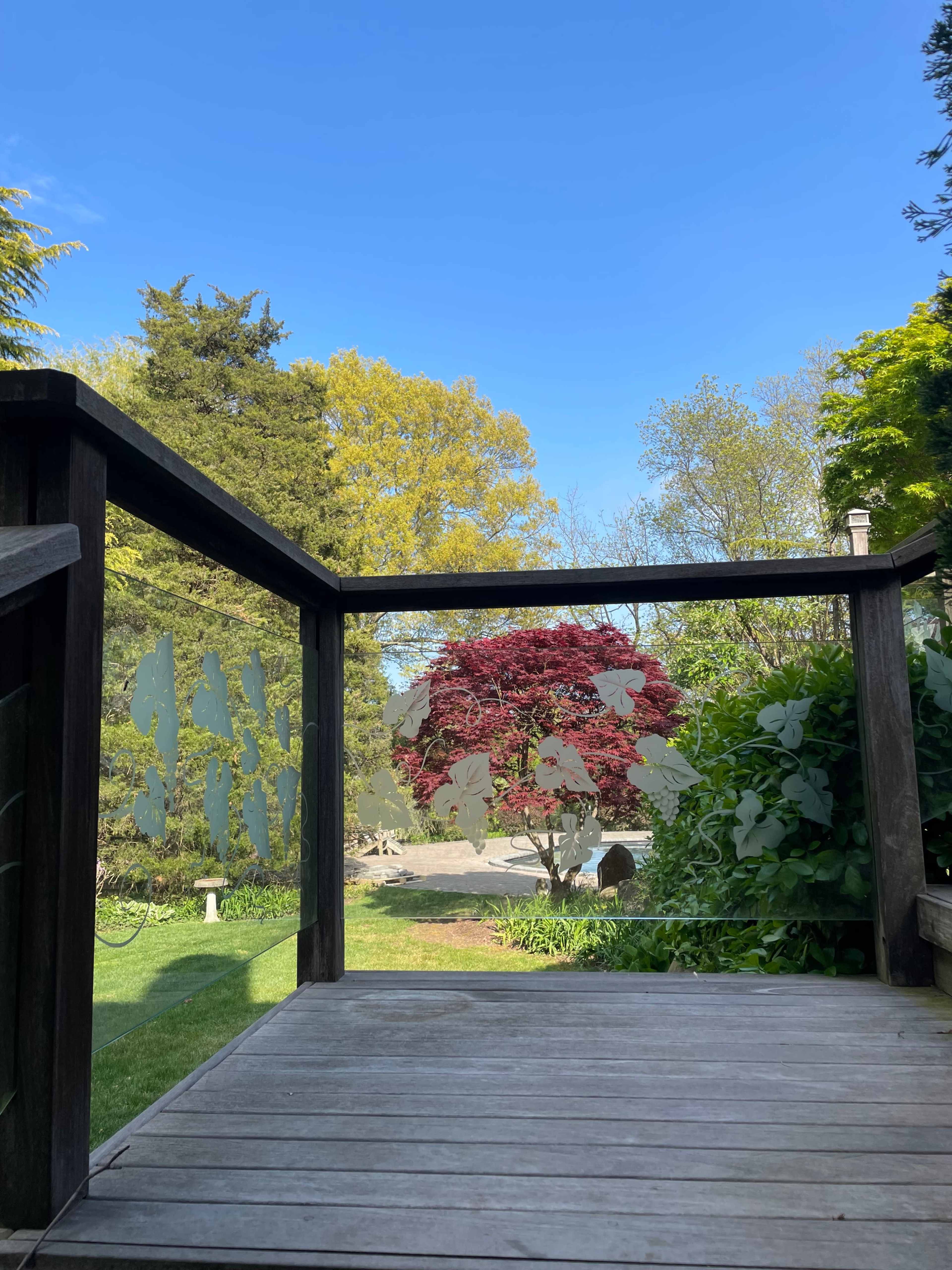 A wooden deck with a glass railing overlooks a well-maintained garden featuring a vibrant red tree and greenery under a clear blue sky.