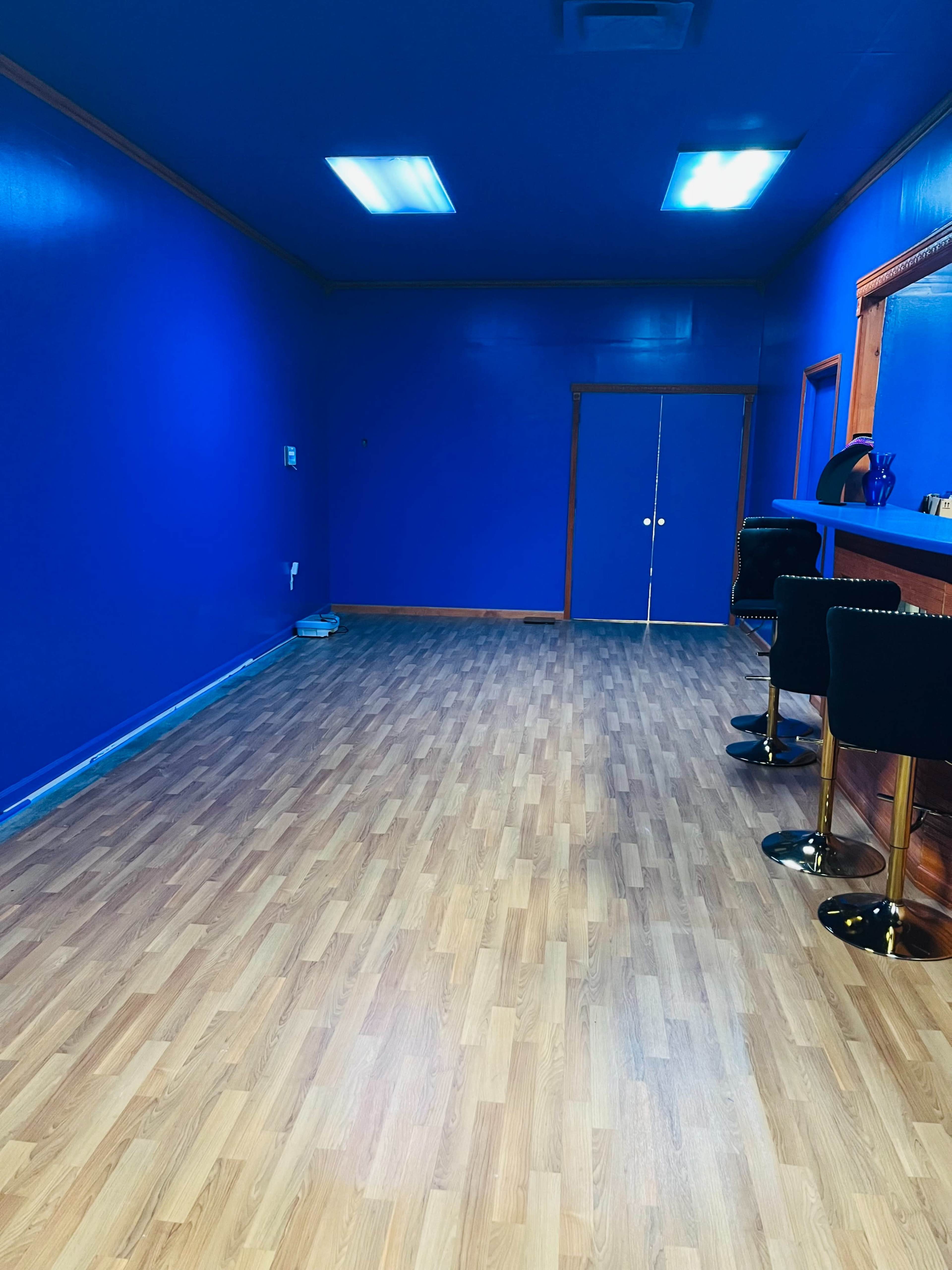 The image shows an empty room with blue walls and wooden flooring, featuring a bar area with two black stools and a door in the background.