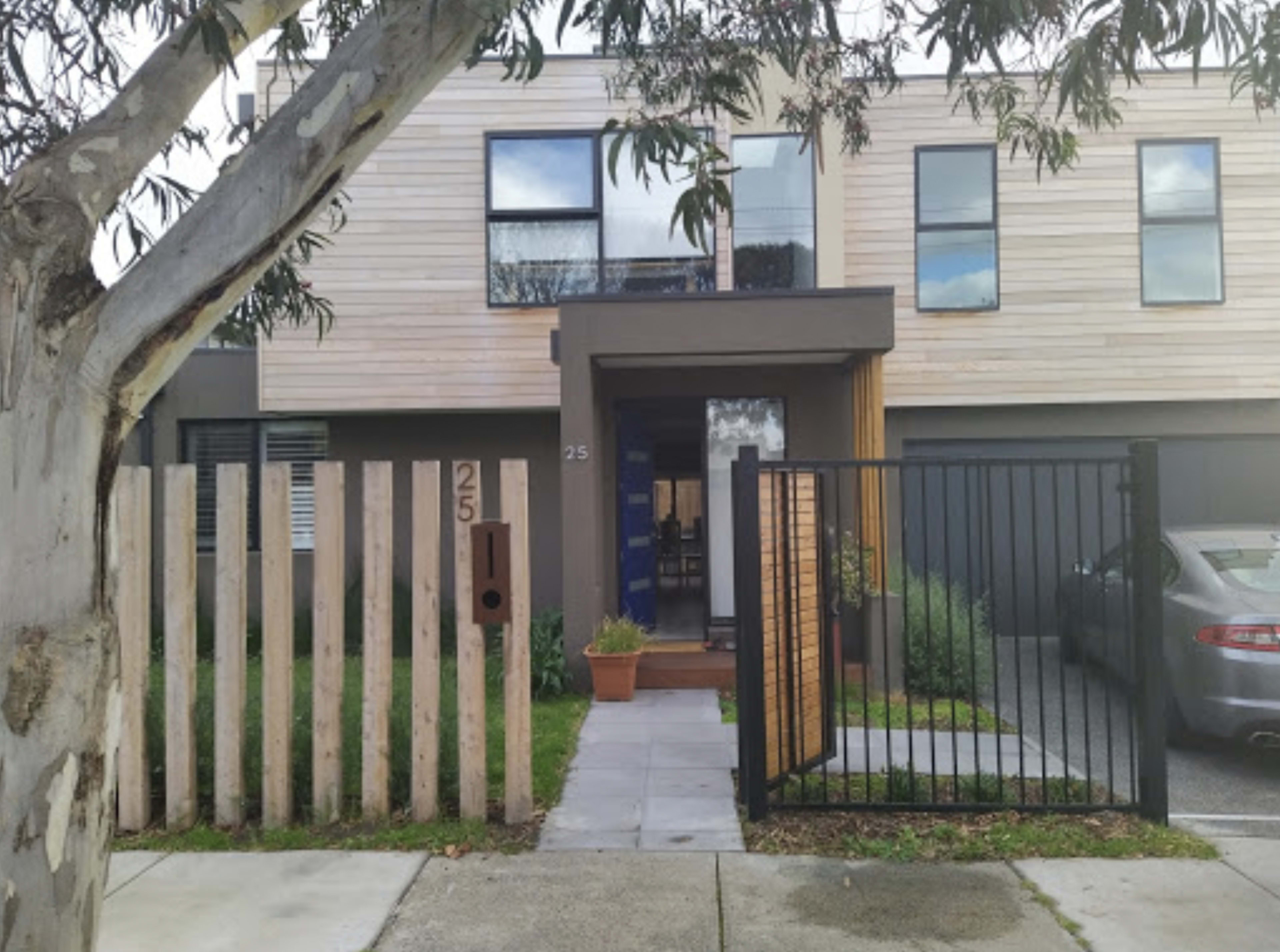 The image shows a modern two-story house with wooden and gray paneling, a front garden, and a gated entrance.