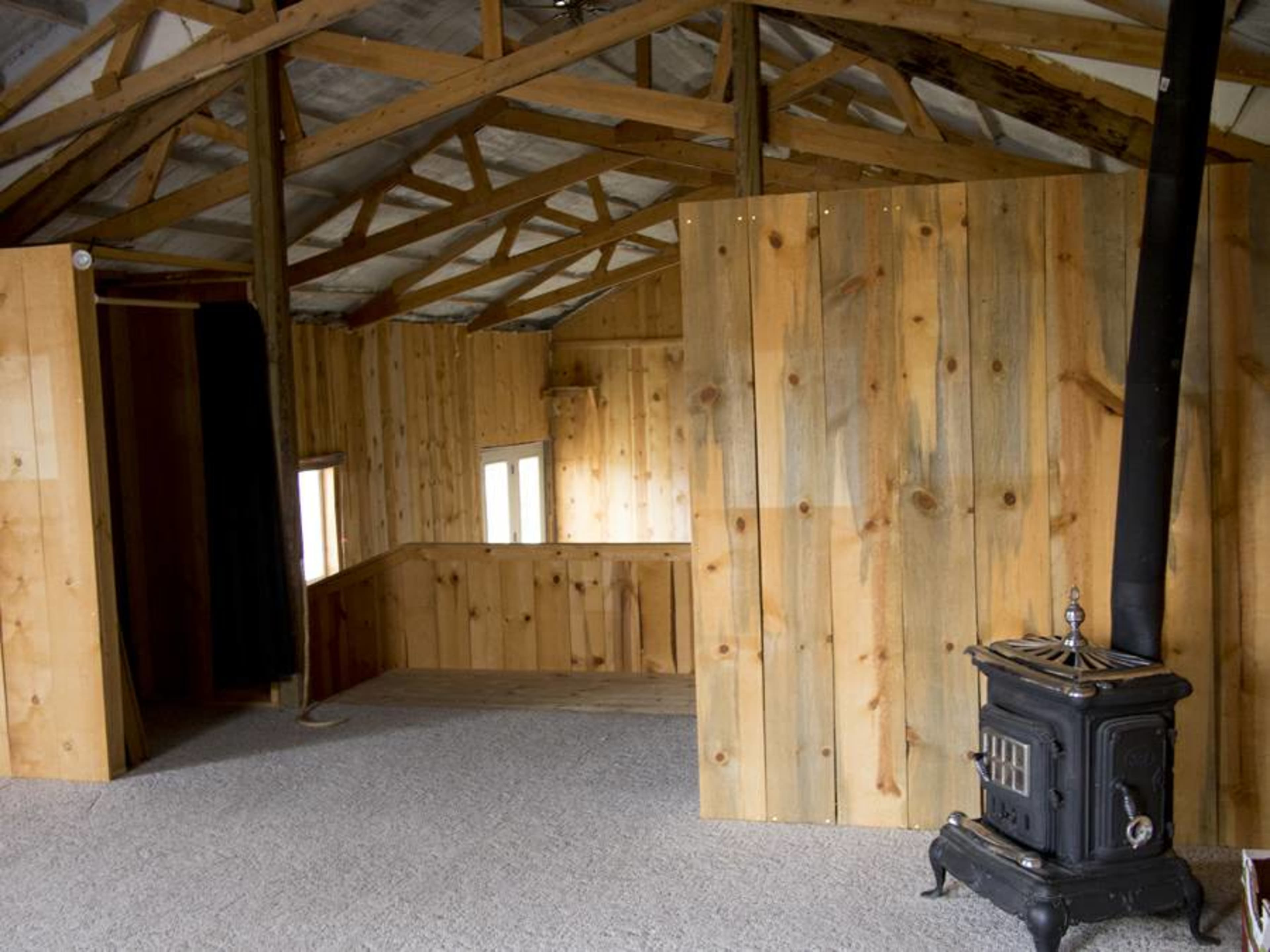 The interior of a cabin features wooden walls, a sloped ceiling with exposed beams, a wood stove, and a carpeted floor.