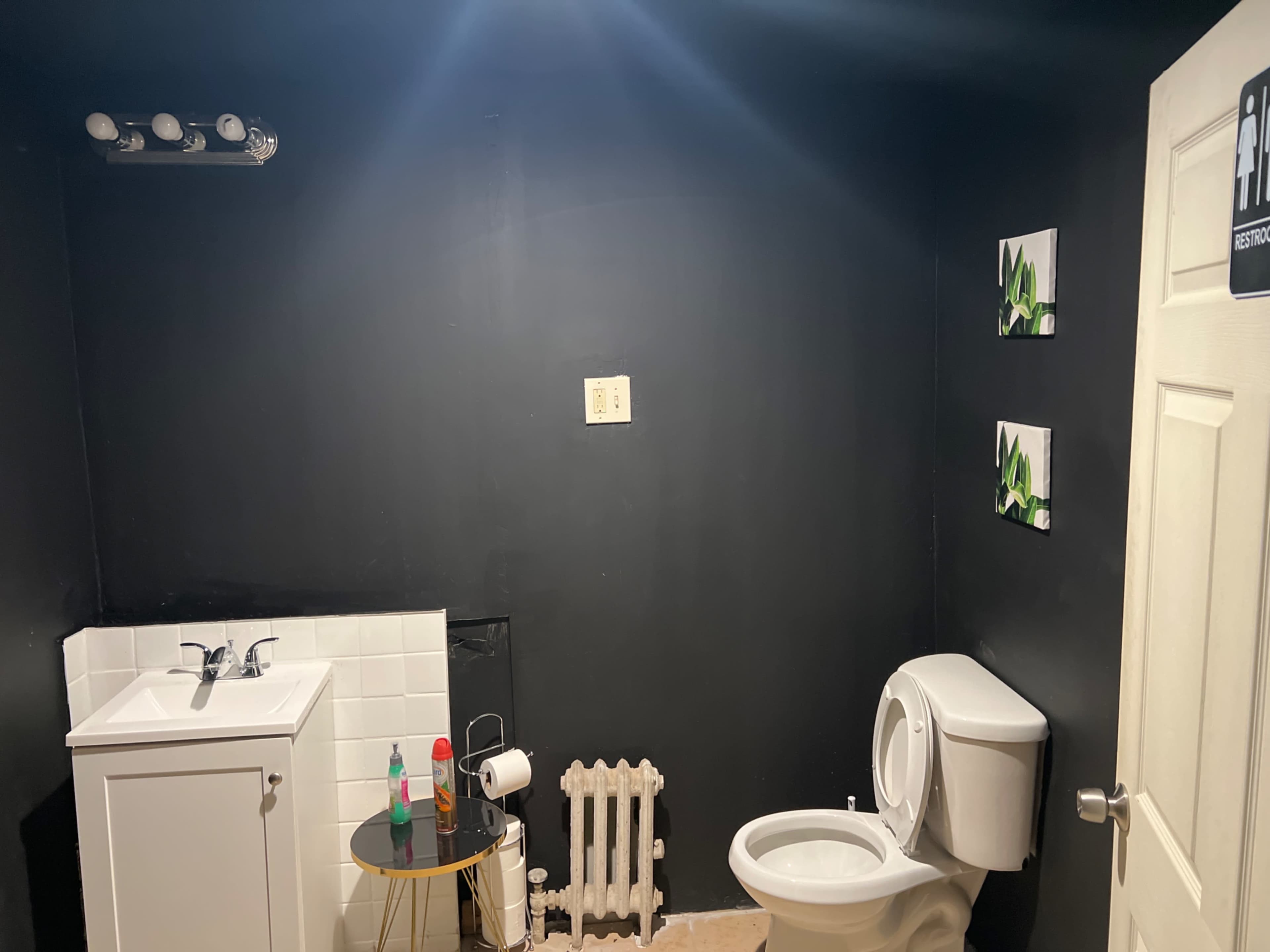 A small bathroom with a black-painted wall, featuring a toilet, a sink, a radiator, and two framed decorations.