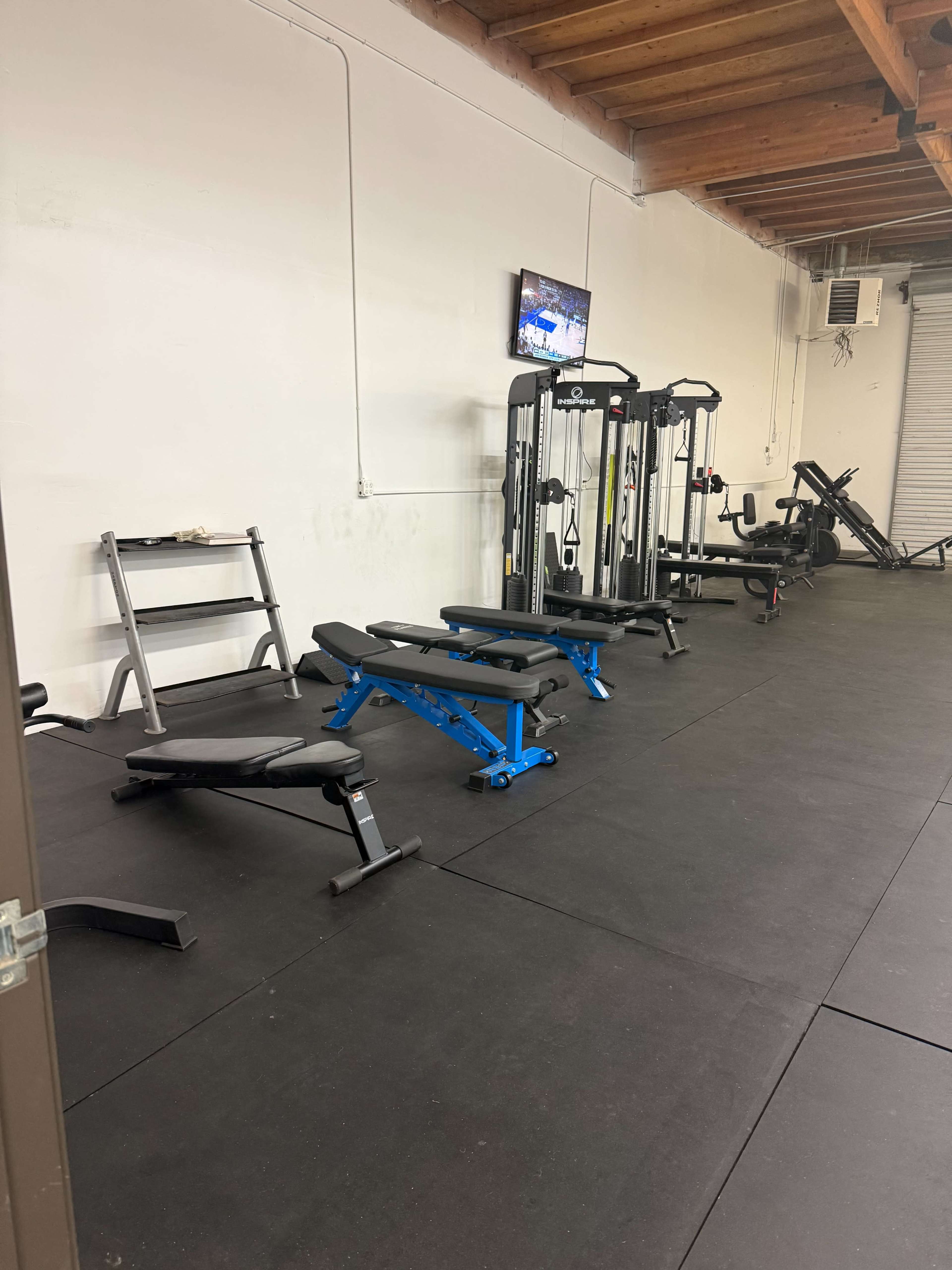 The image shows a sparsely equipped gym area featuring benches, weight machines, and a television mounted on the wall.