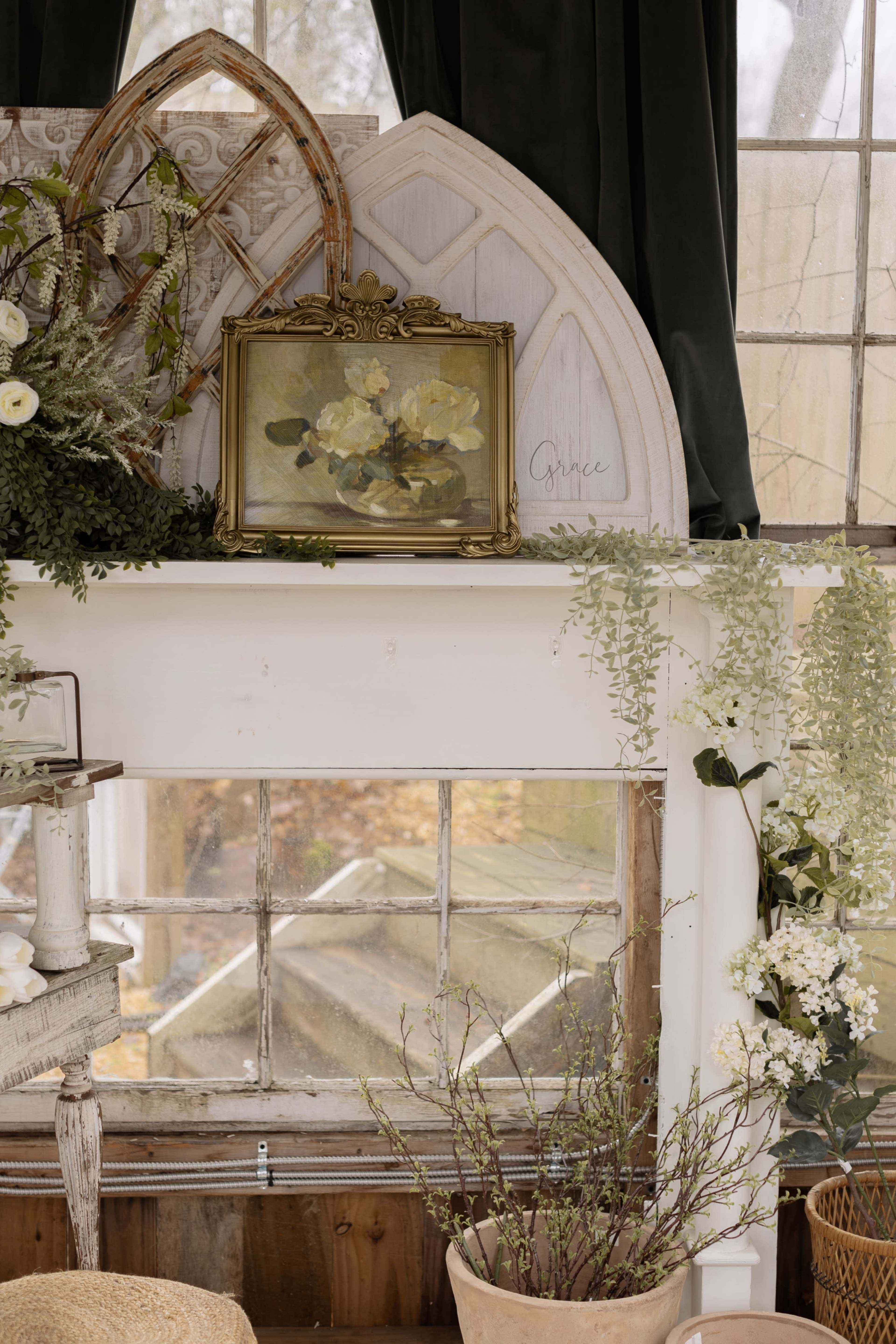 The image shows a room with a decorative mantel featuring a framed painting, various plants, and decorative elements against a backdrop of large windows.