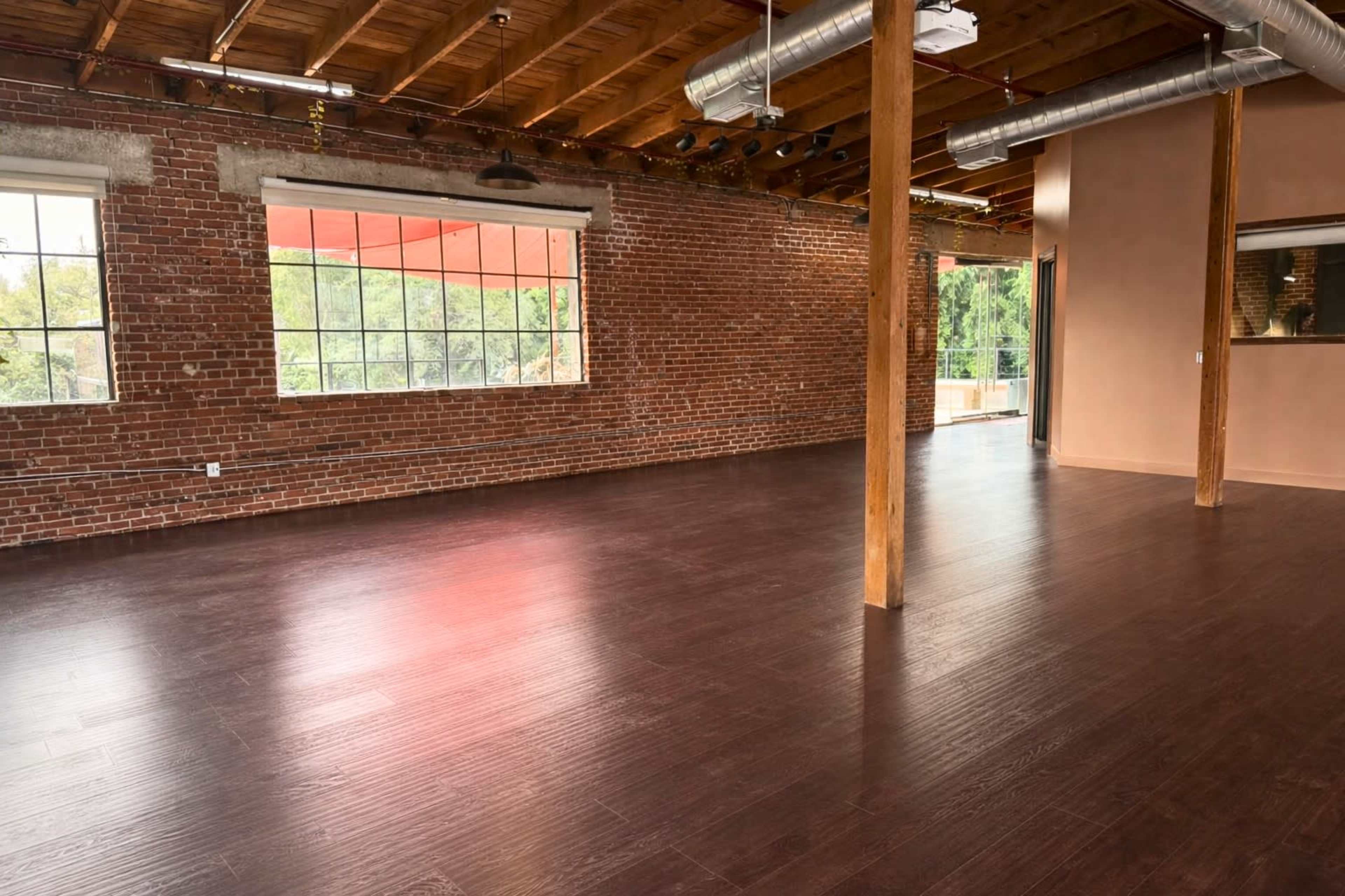 The image shows a spacious room with exposed brick walls, wooden beams, and large windows, featuring a polished hardwood floor.