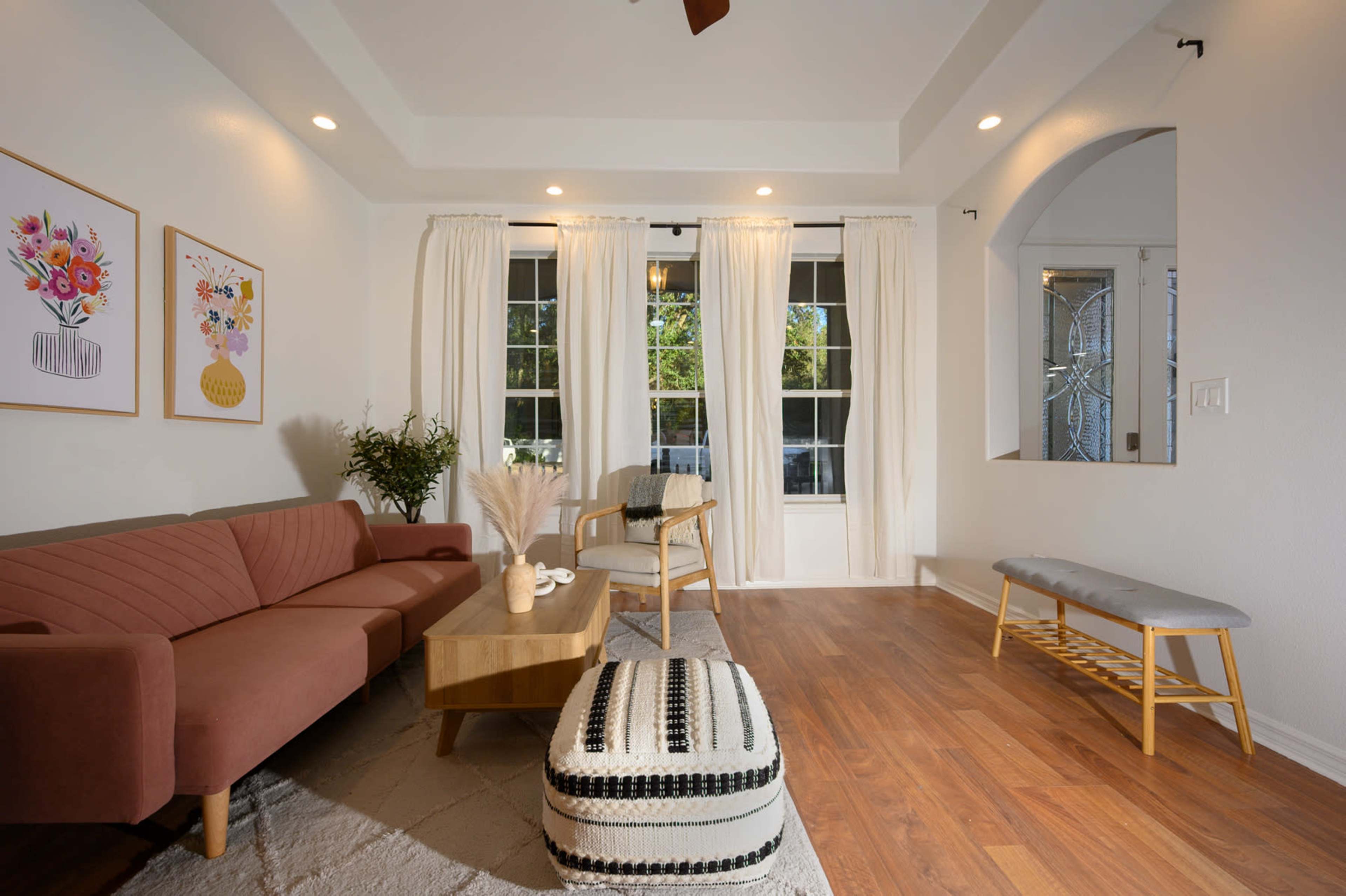 A modern living room features a pink sofa, a wooden coffee table, and large windows with sheer curtains.