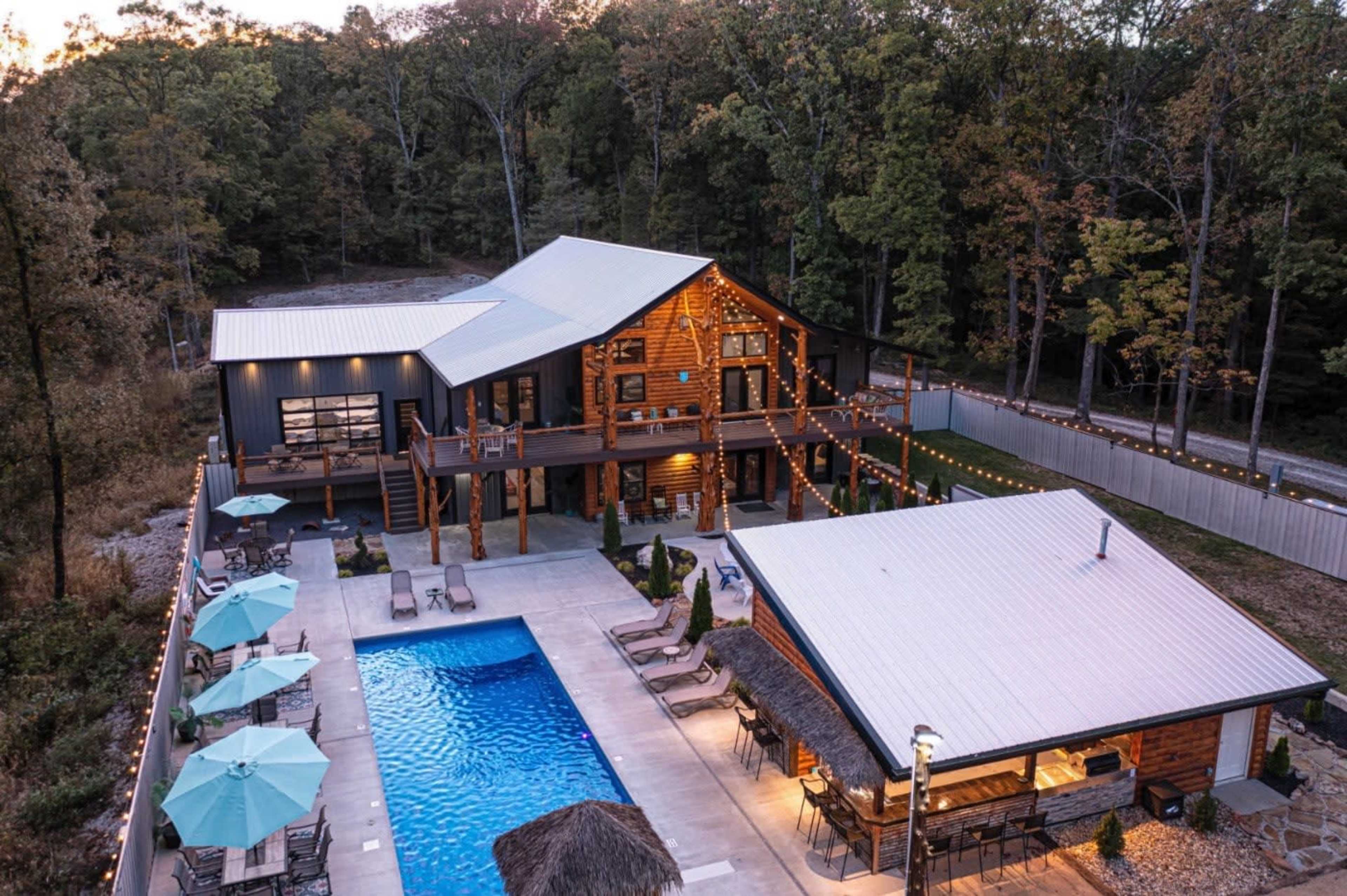 A modern lodge-style home with a large pool and surrounding outdoor seating area, set in a wooded environment.
