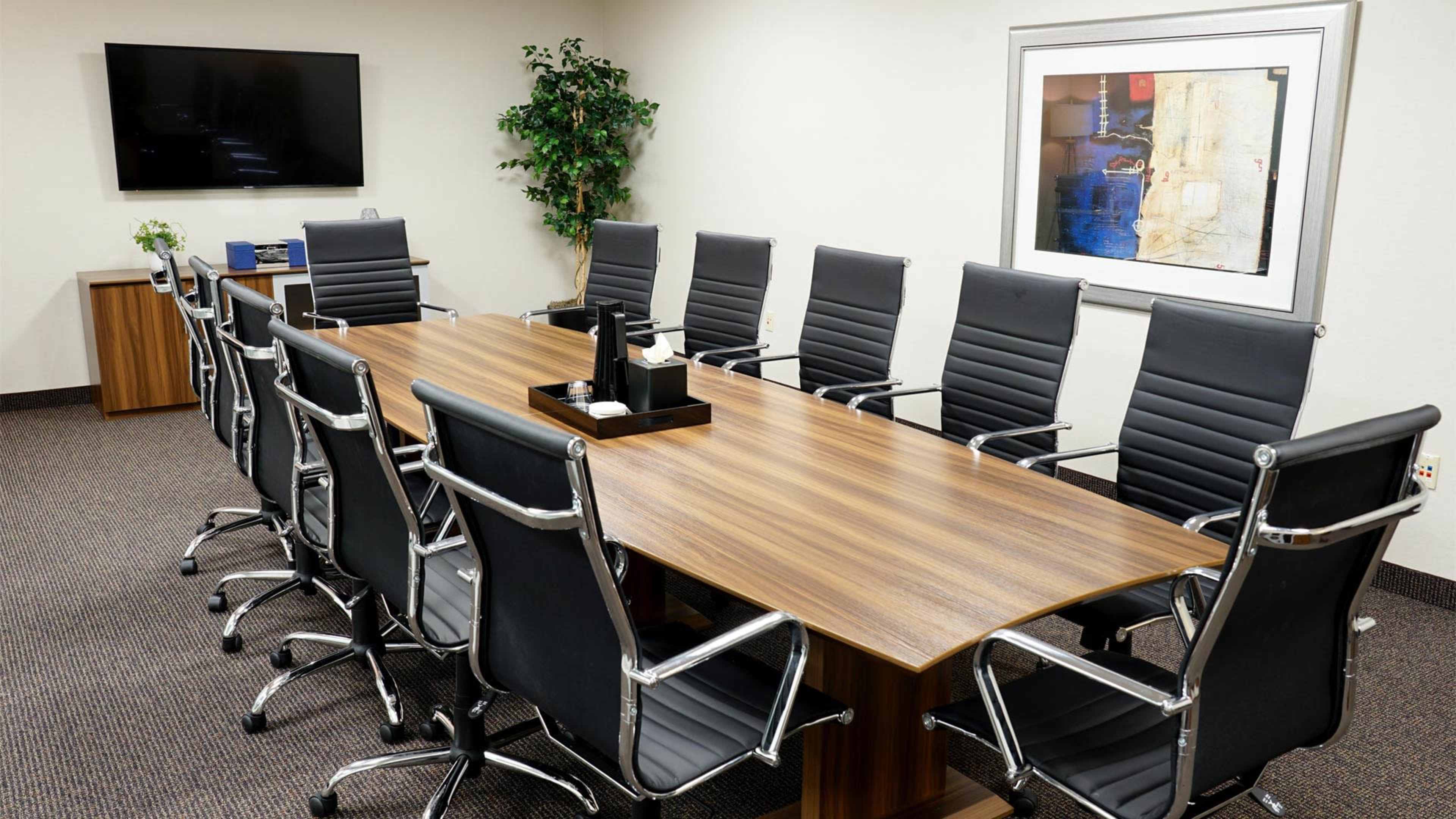A modern conference room features a long wooden table surrounded by black ergonomic chairs, with a television mounted on the wall and a framed artwork.