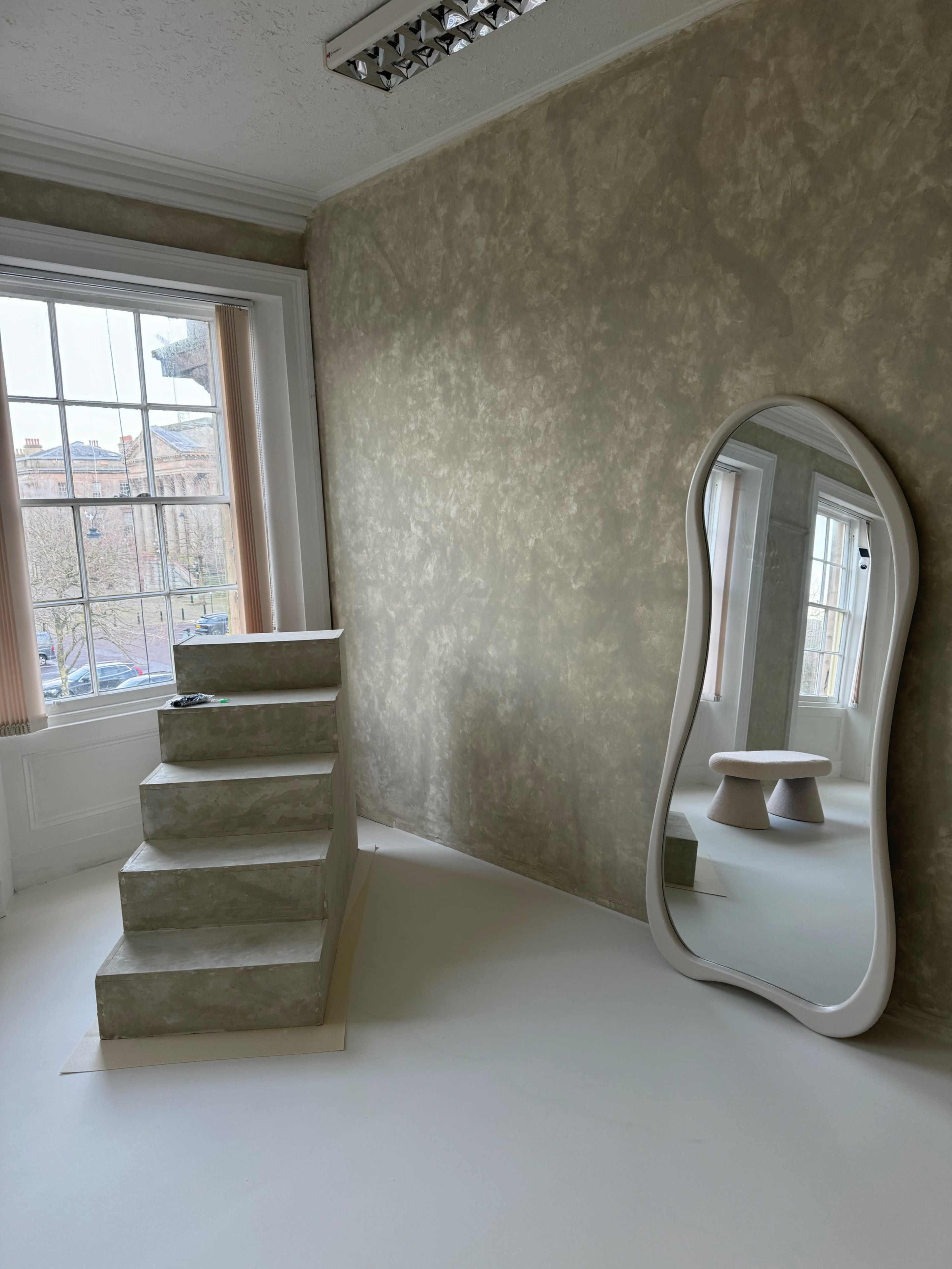 The image shows a modern interior space featuring a curved mirror, a small round table, and a set of steps against a textured wall, with large windows revealing a view outside.