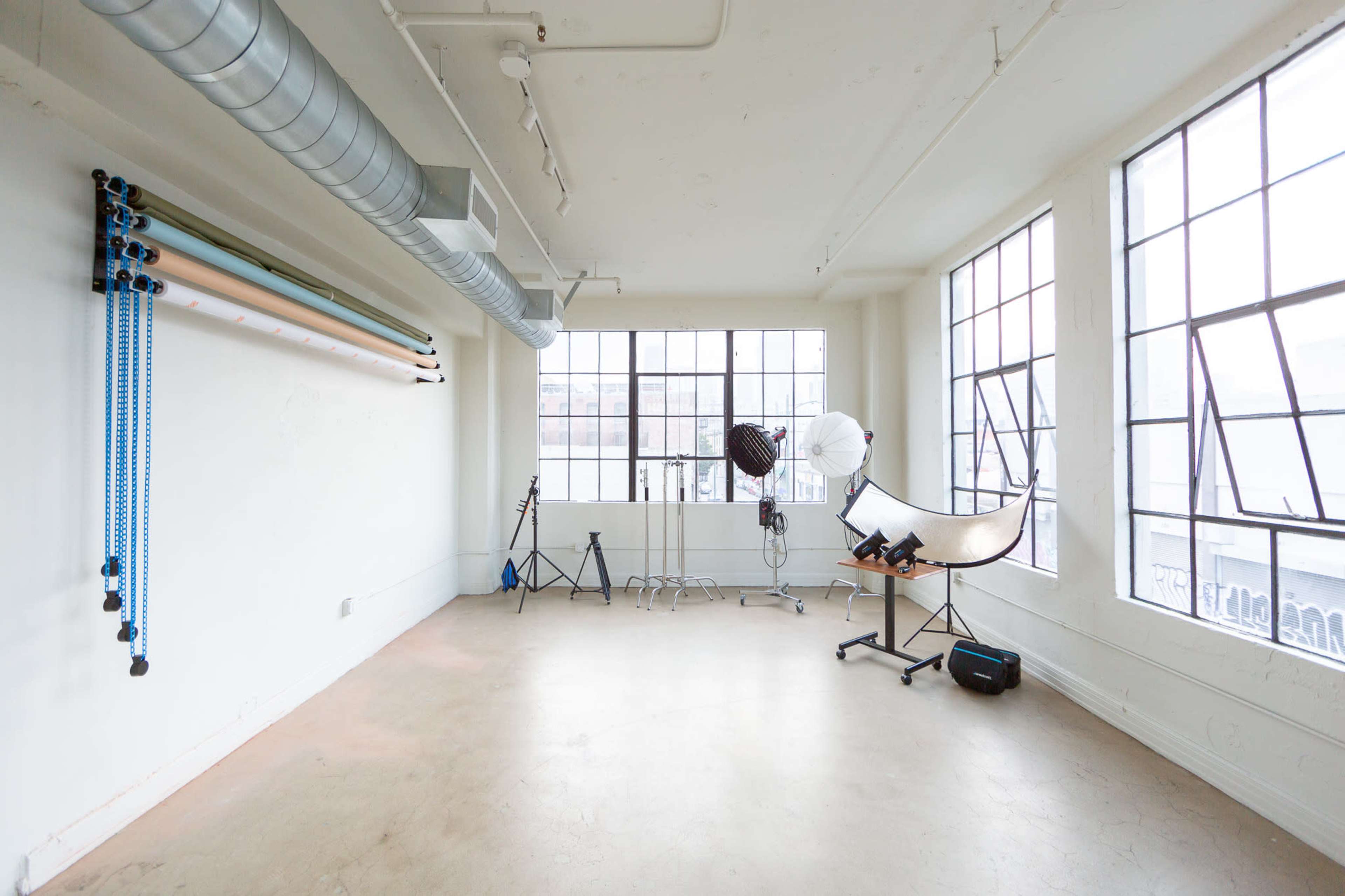 A spacious studio with large windows, studio lighting equipment, and a backdrop support system neatly arranged along the walls.