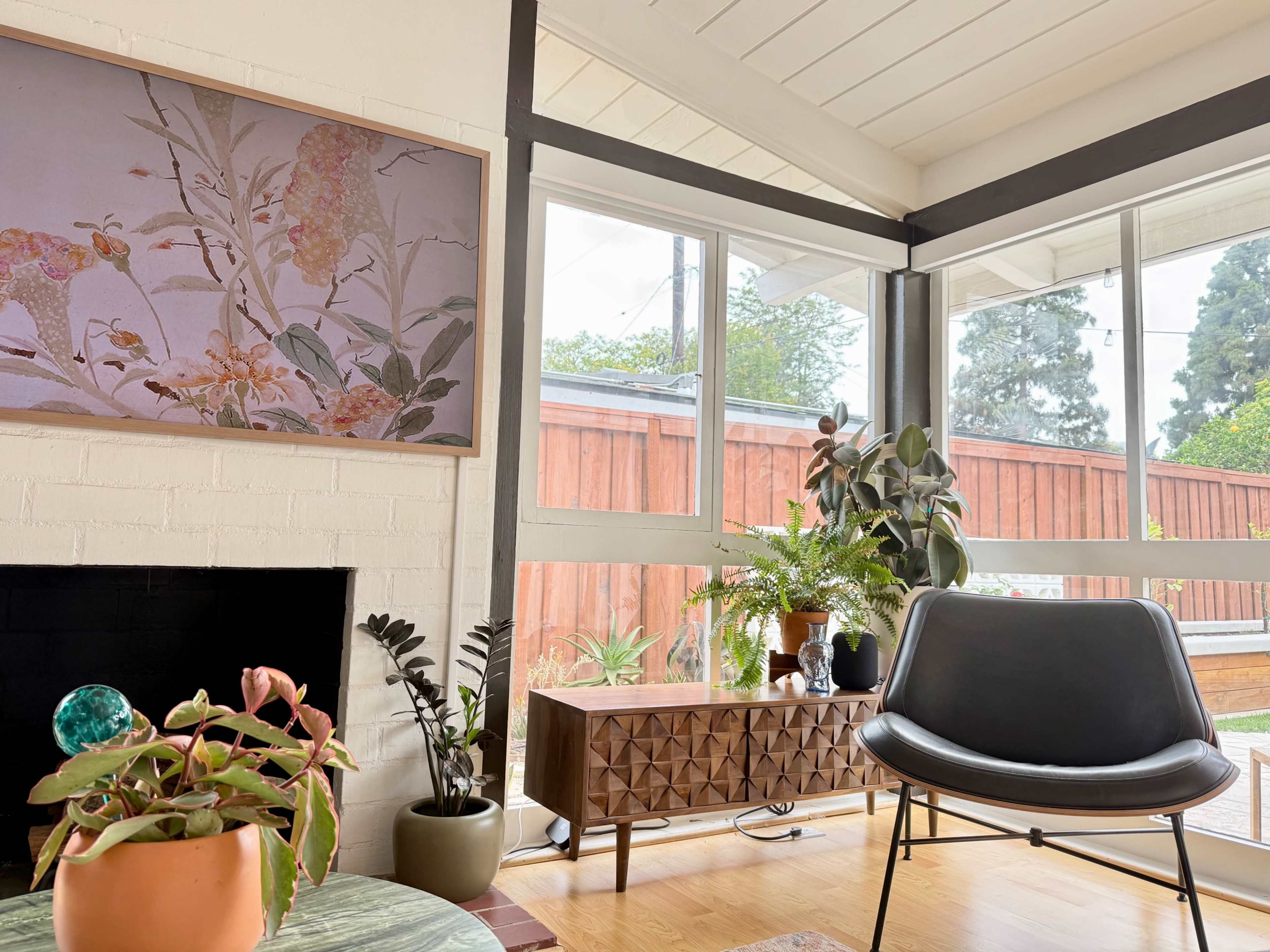 A cozy living room features a large floral artwork on the wall, a modern chair, a wooden sideboard, and plants placed throughout the space.