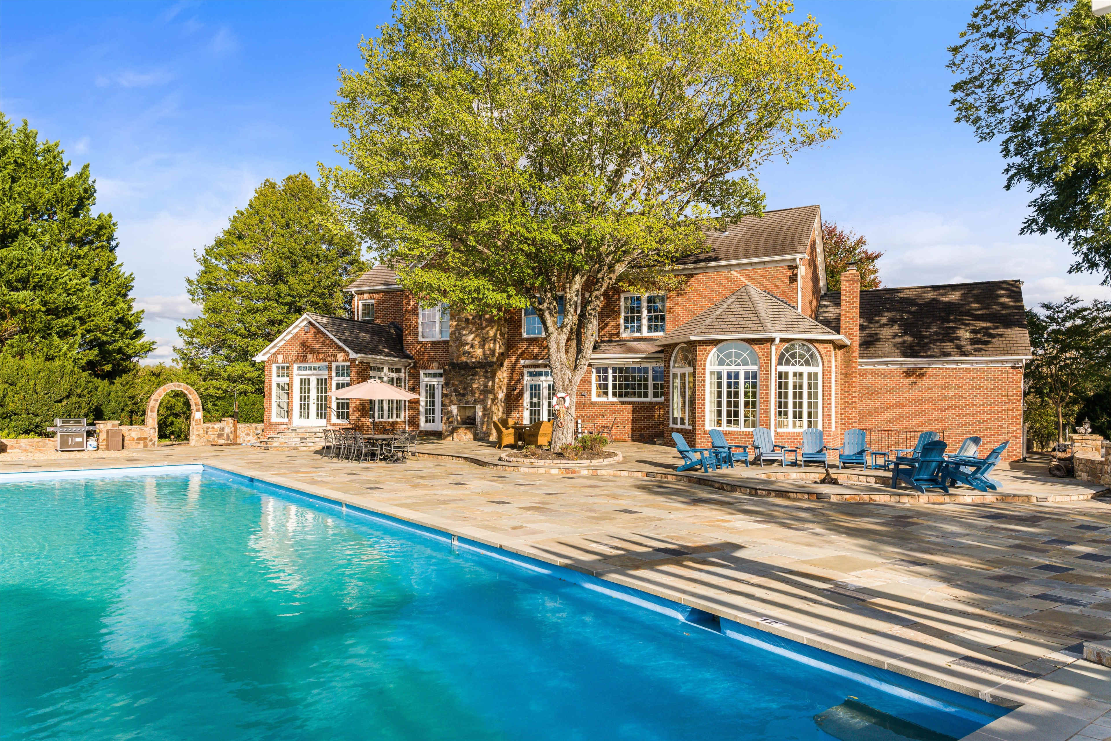 The image shows a large brick house with multiple windows and a swimming pool in the foreground, surrounded by a stone patio and outdoor seating.