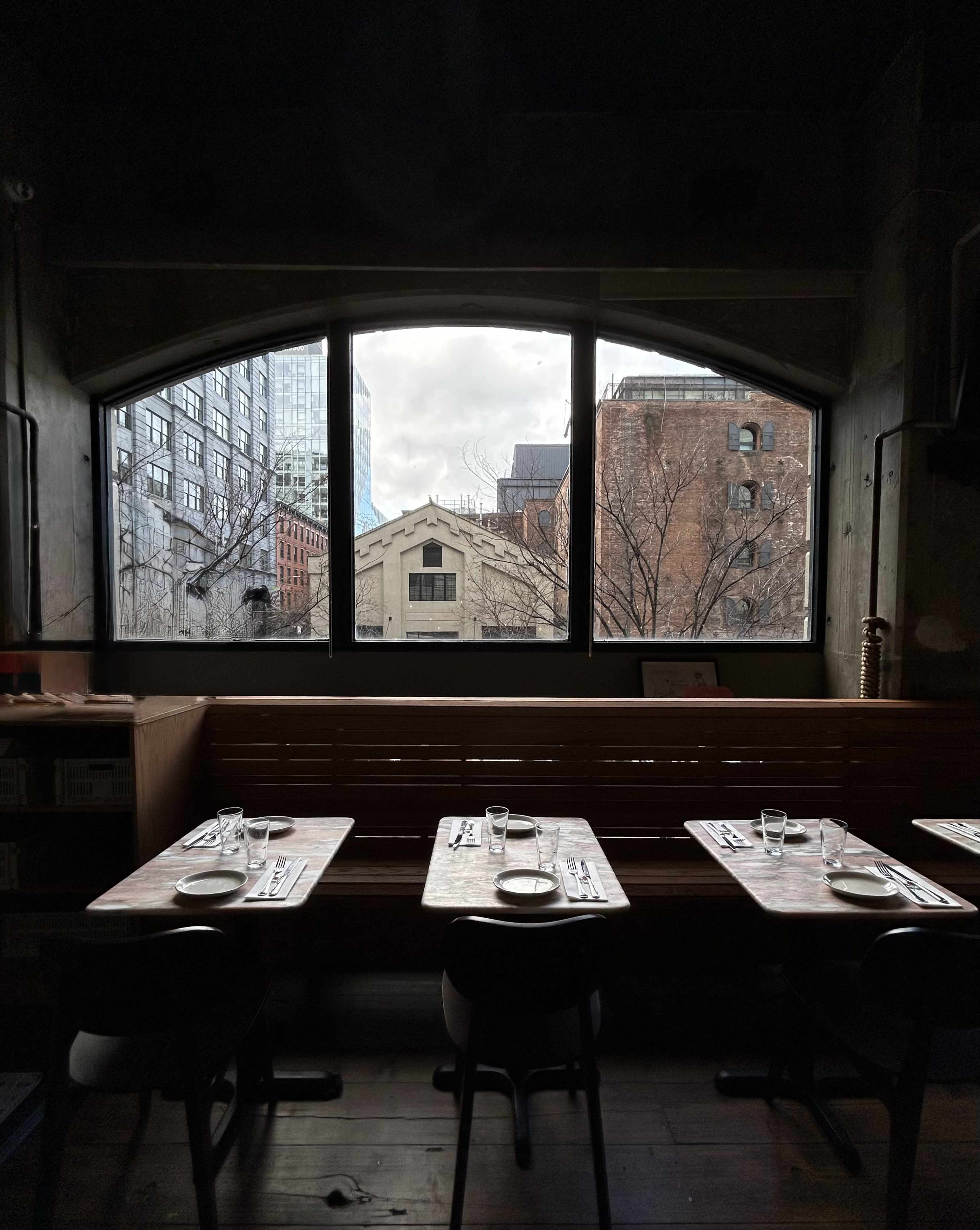 A restaurant interior features empty tables set for dining, facing a large window that overlooks a cityscape with bare trees and buildings.