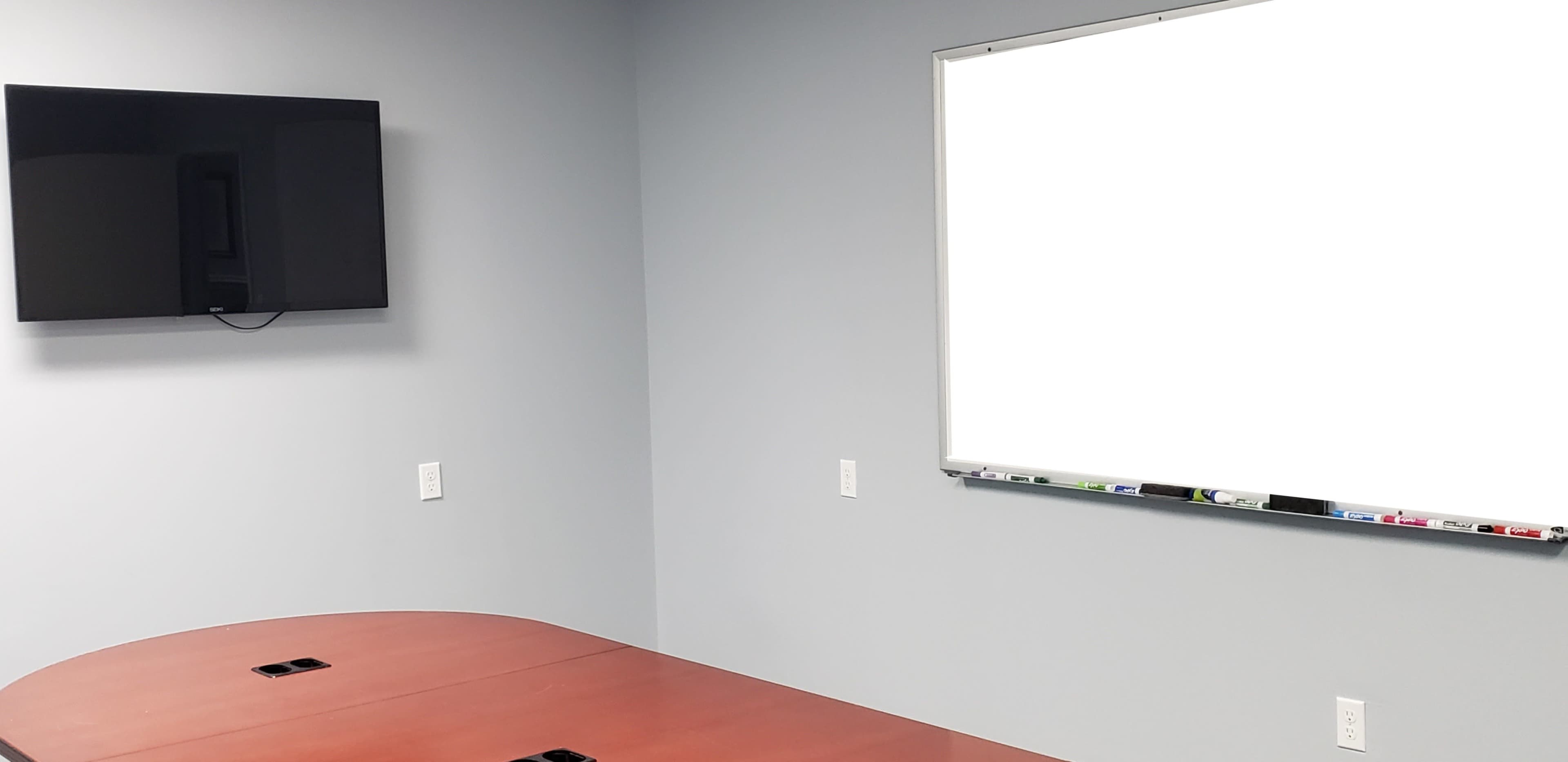 A meeting room with a brown table, a wall-mounted television, and a blank whiteboard against a gray wall.