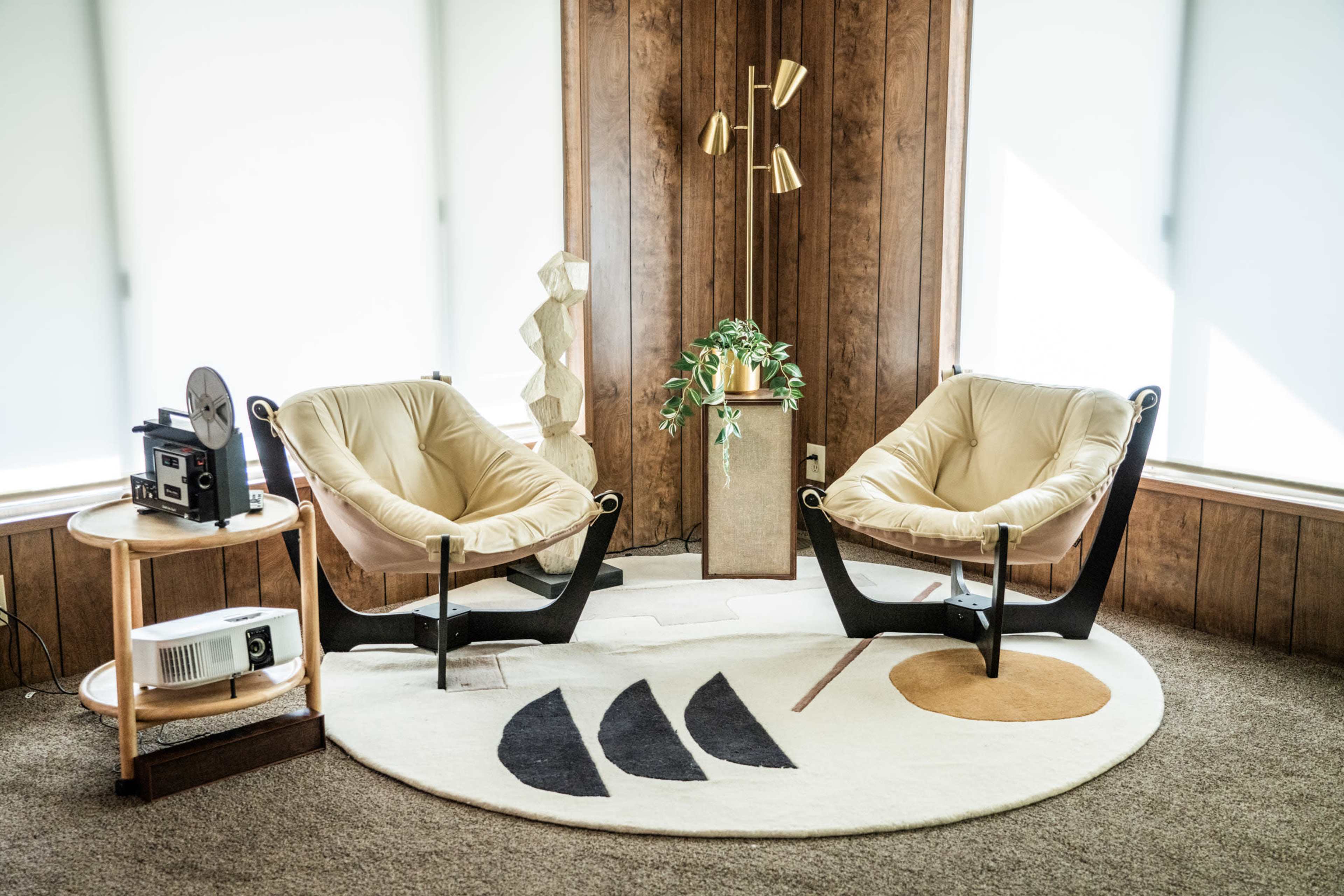 The image features two modern armchairs on a circular rug in a room with wooden paneling and large windows, accompanied by a plant and a film projector on a side table.