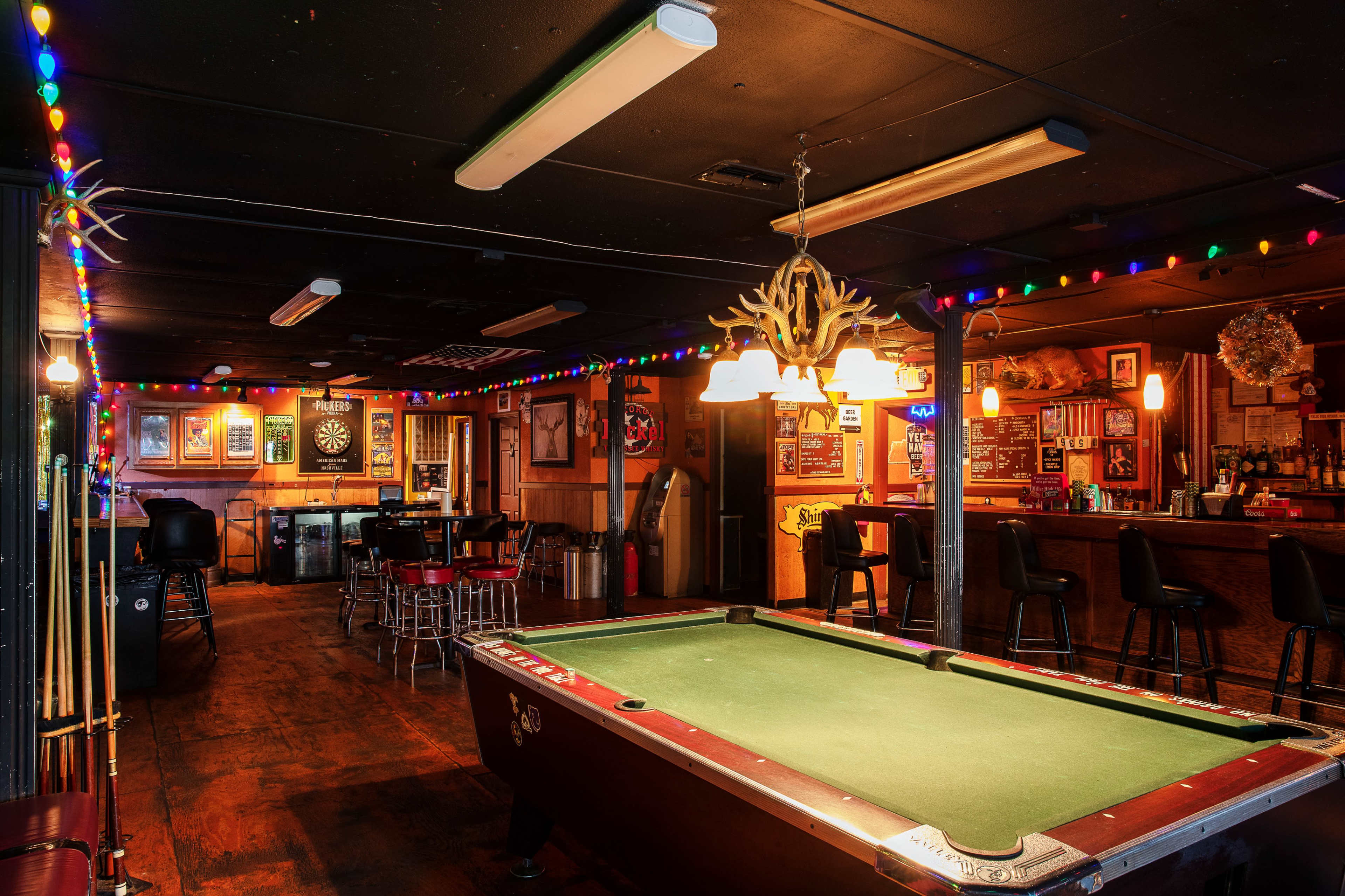 The image shows a dimly lit barroom featuring a pool table in the foreground, a bar counter in the back, and colorful string lights decorating the ceiling.