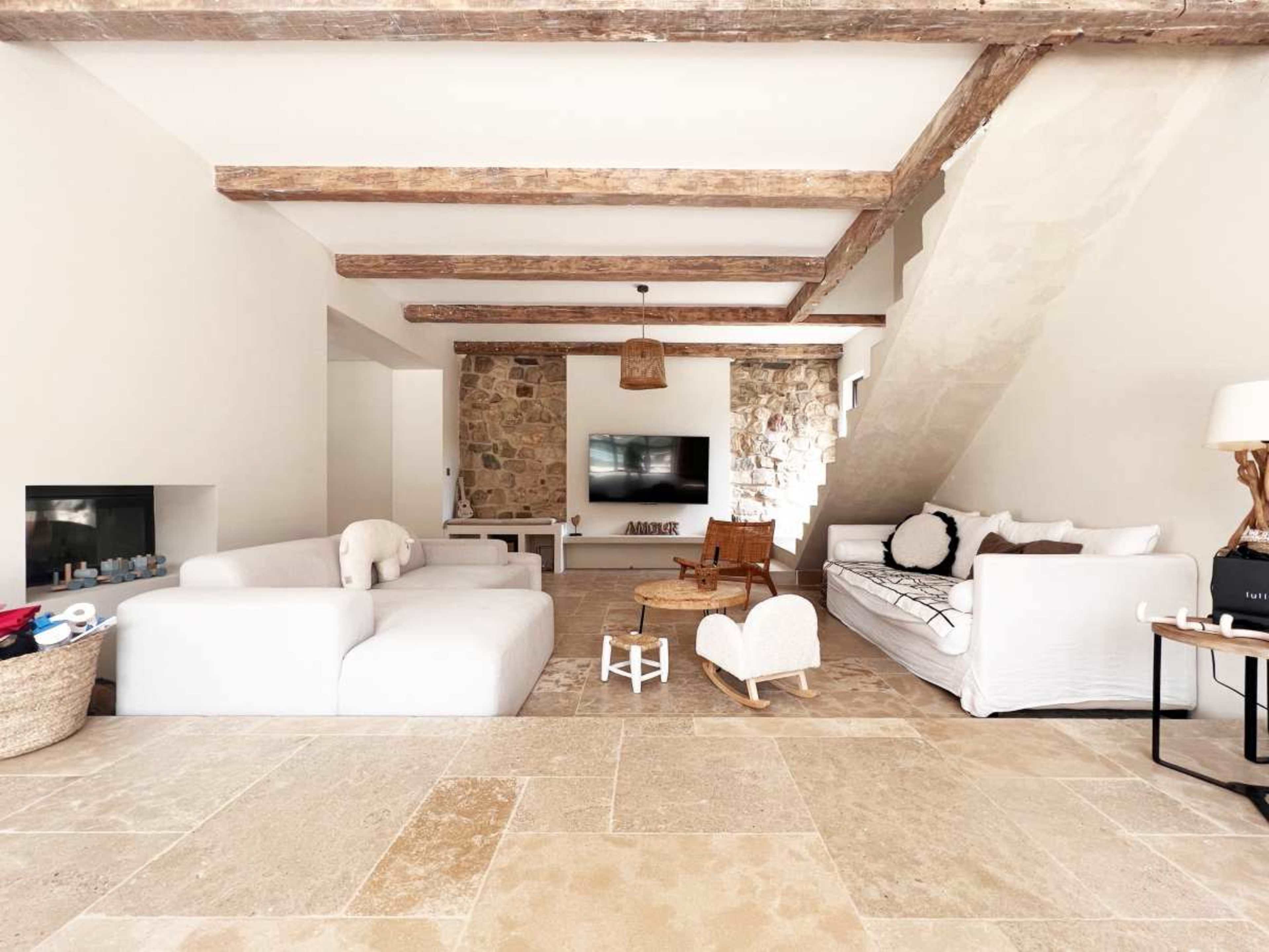 The living room features a minimalist design with a light-colored sectional sofa, a stone wall with a TV, and wooden beams overhead.