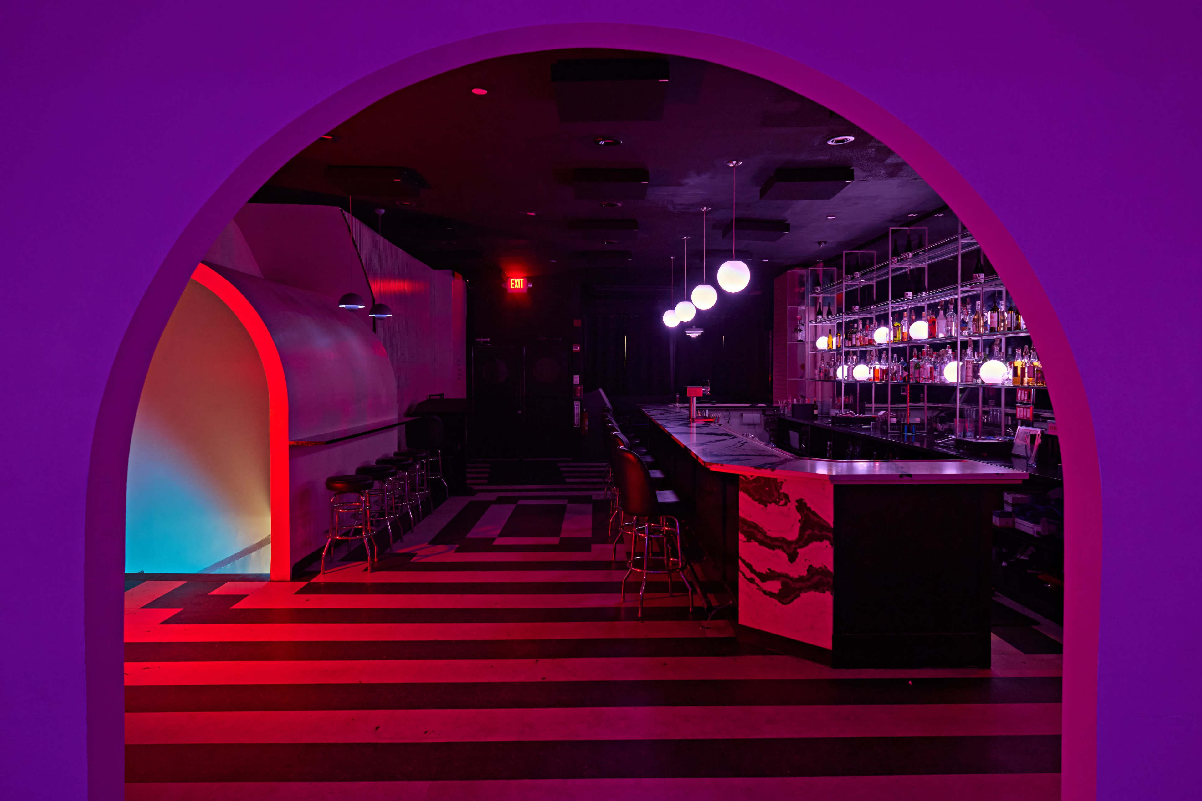 A dimly lit bar scene with a striped floor and colorful lighting, featuring a long bar counter and a row of spherical pendant lights.