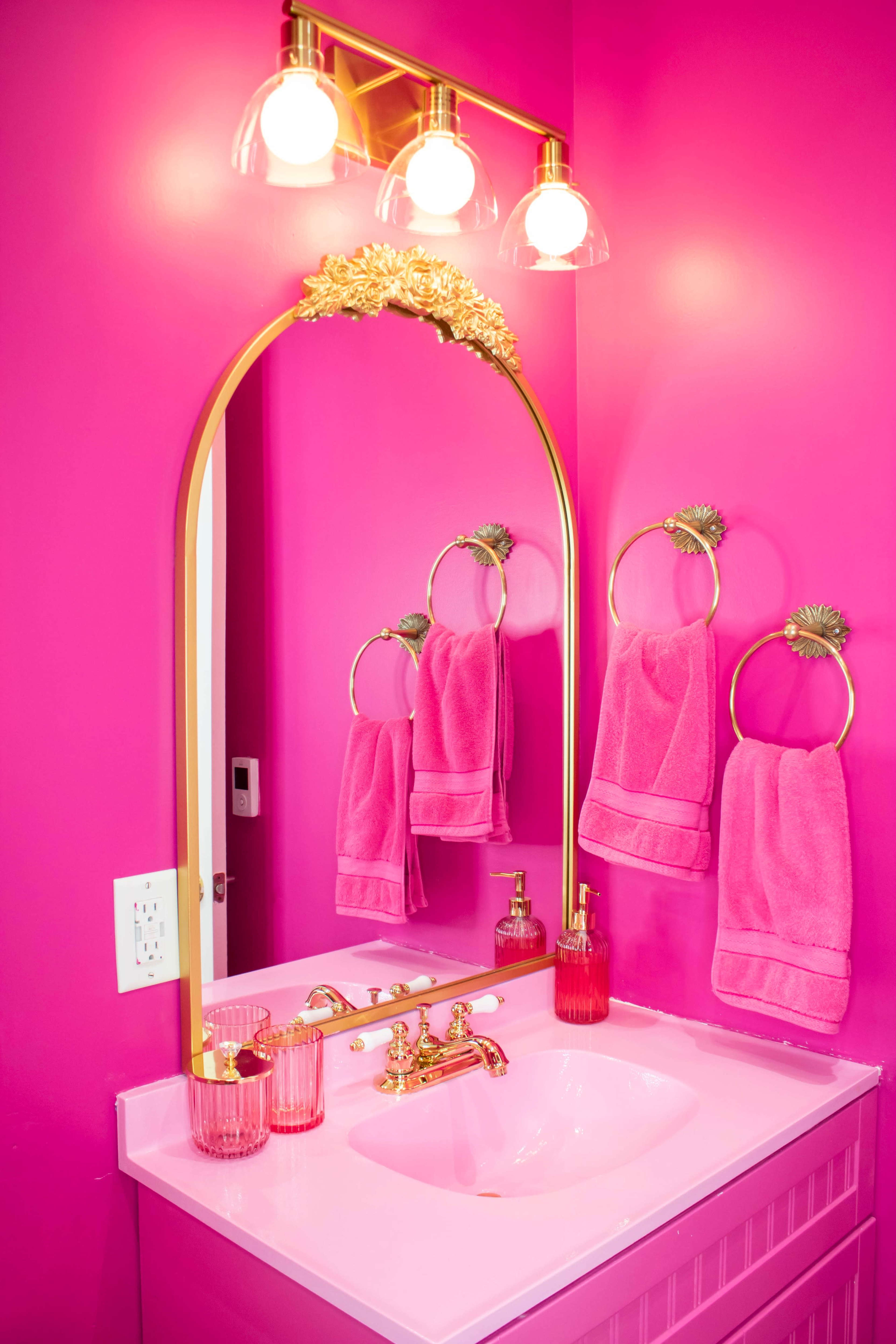 A pink bathroom features a gold-framed mirror, gold fixtures, and matching towels hanging from towel rings.