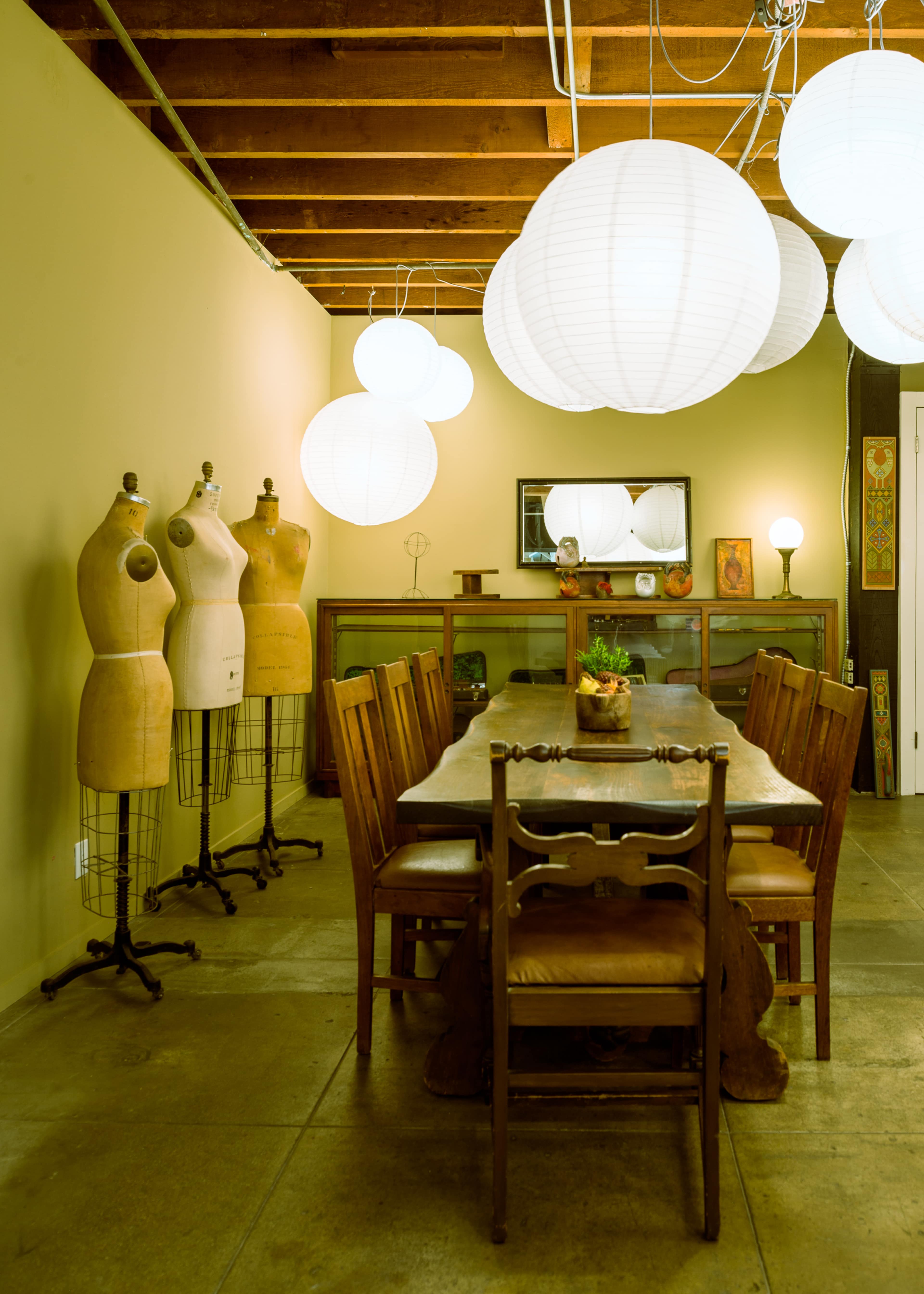 A spacious dining area with a long wooden table surrounded by chairs, mannequins on stands, and paper lanterns hanging overhead.