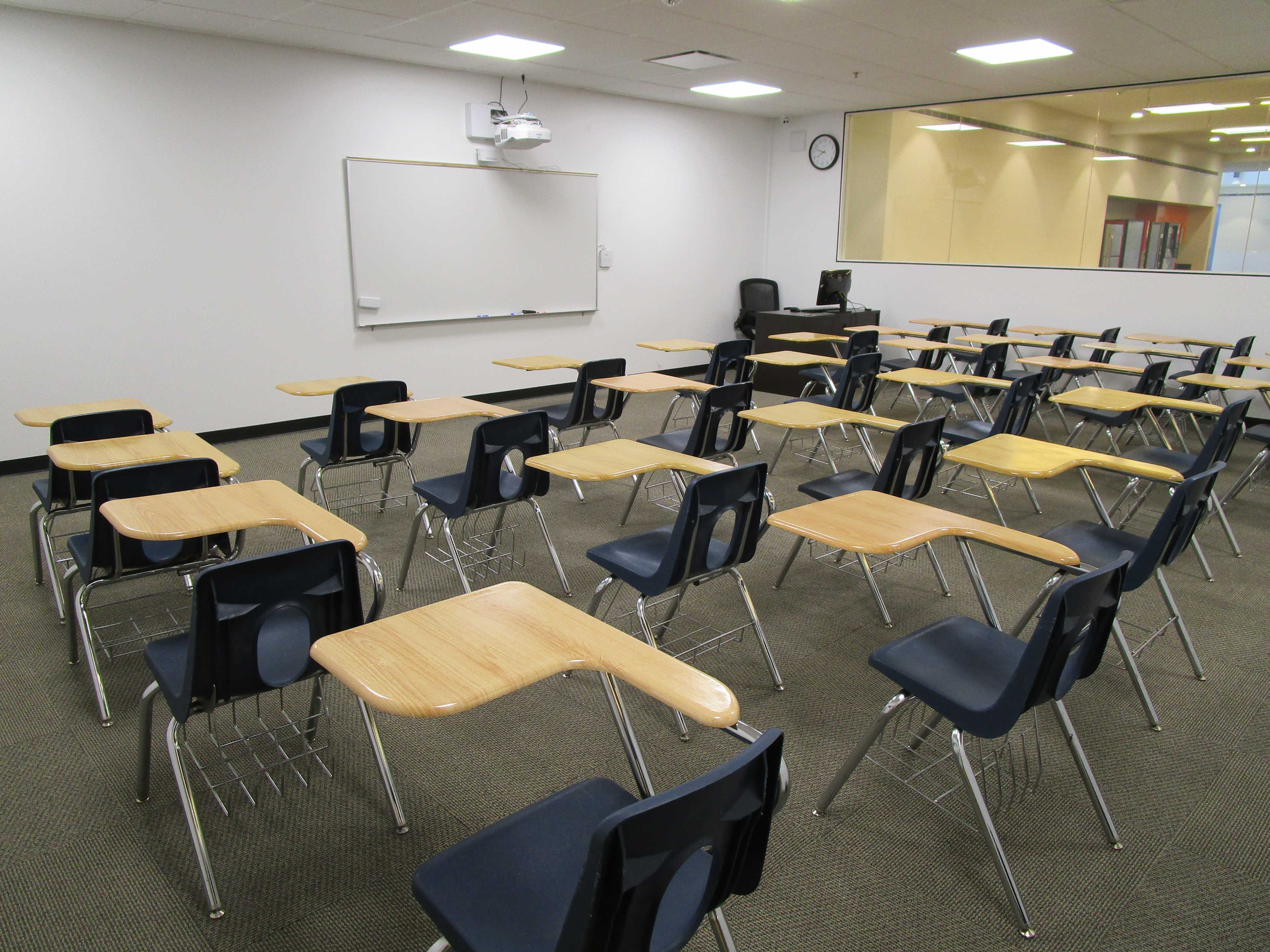 The image shows a classroom arranged with multiple rows of empty desks and chairs, facing a whiteboard.