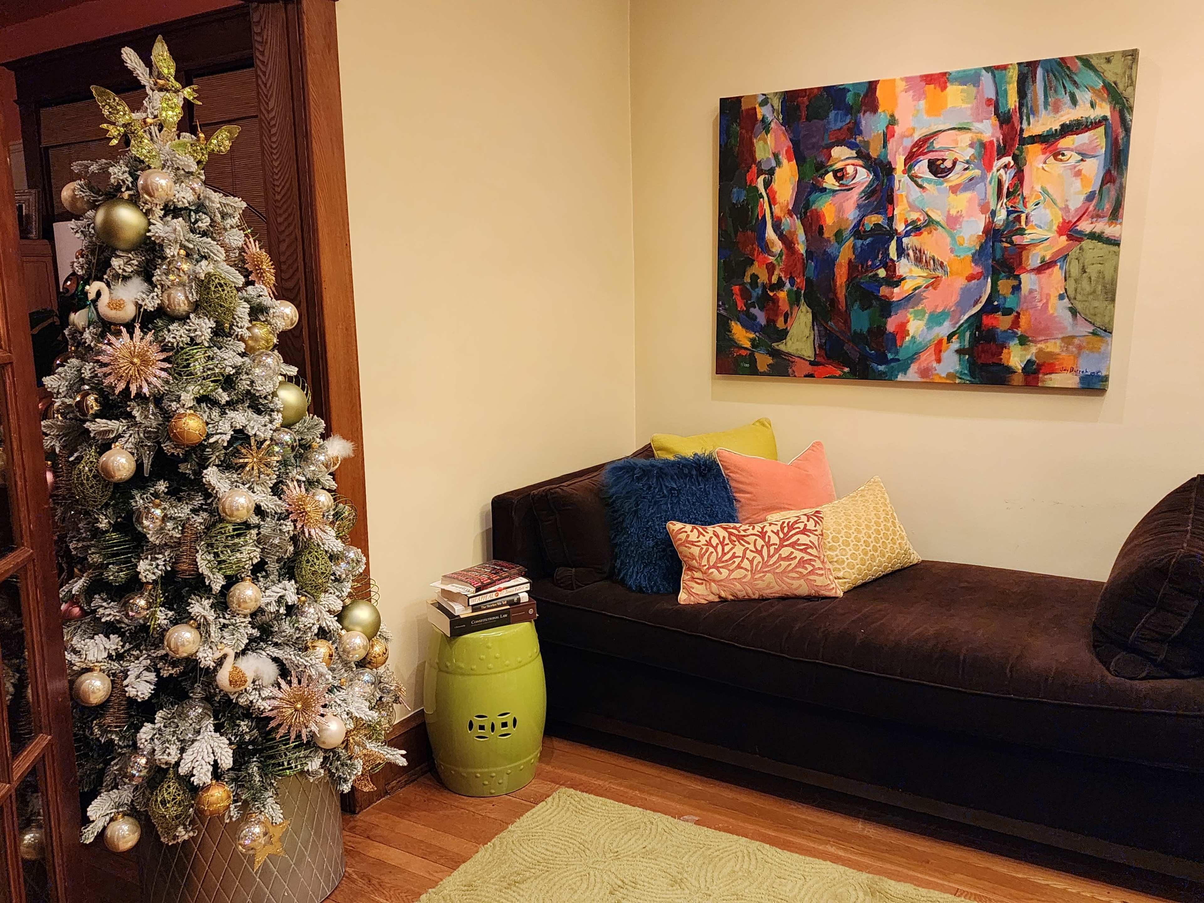 A decorated Christmas tree with metallic ornaments stands beside a dark couch adorned with colorful cushions, while a vibrant abstract painting hangs on the wall above.