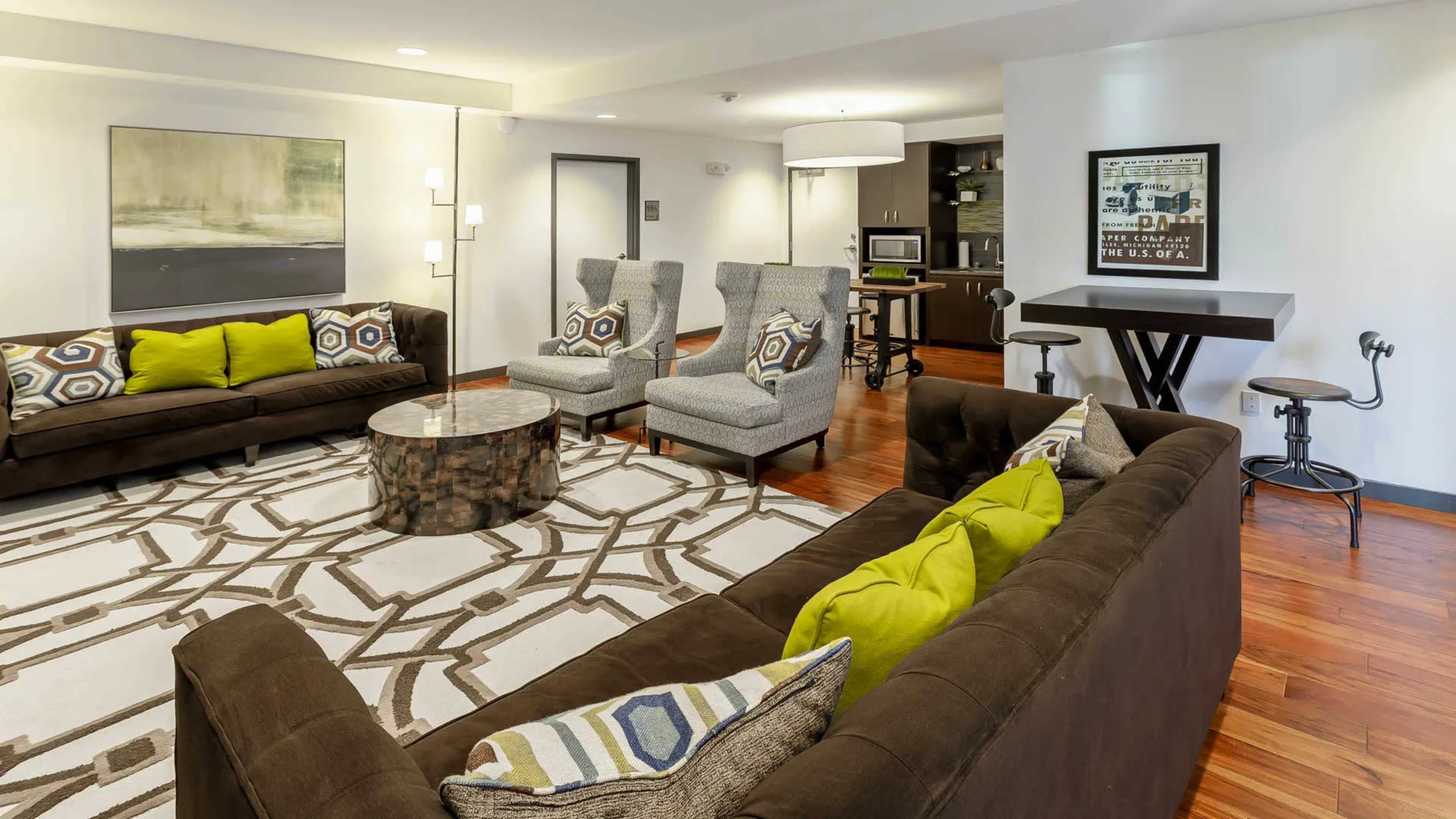 A modern living room furnished with two sofas, two patterned chairs, a circular coffee table, and a rug, with a kitchenette visible in the background.