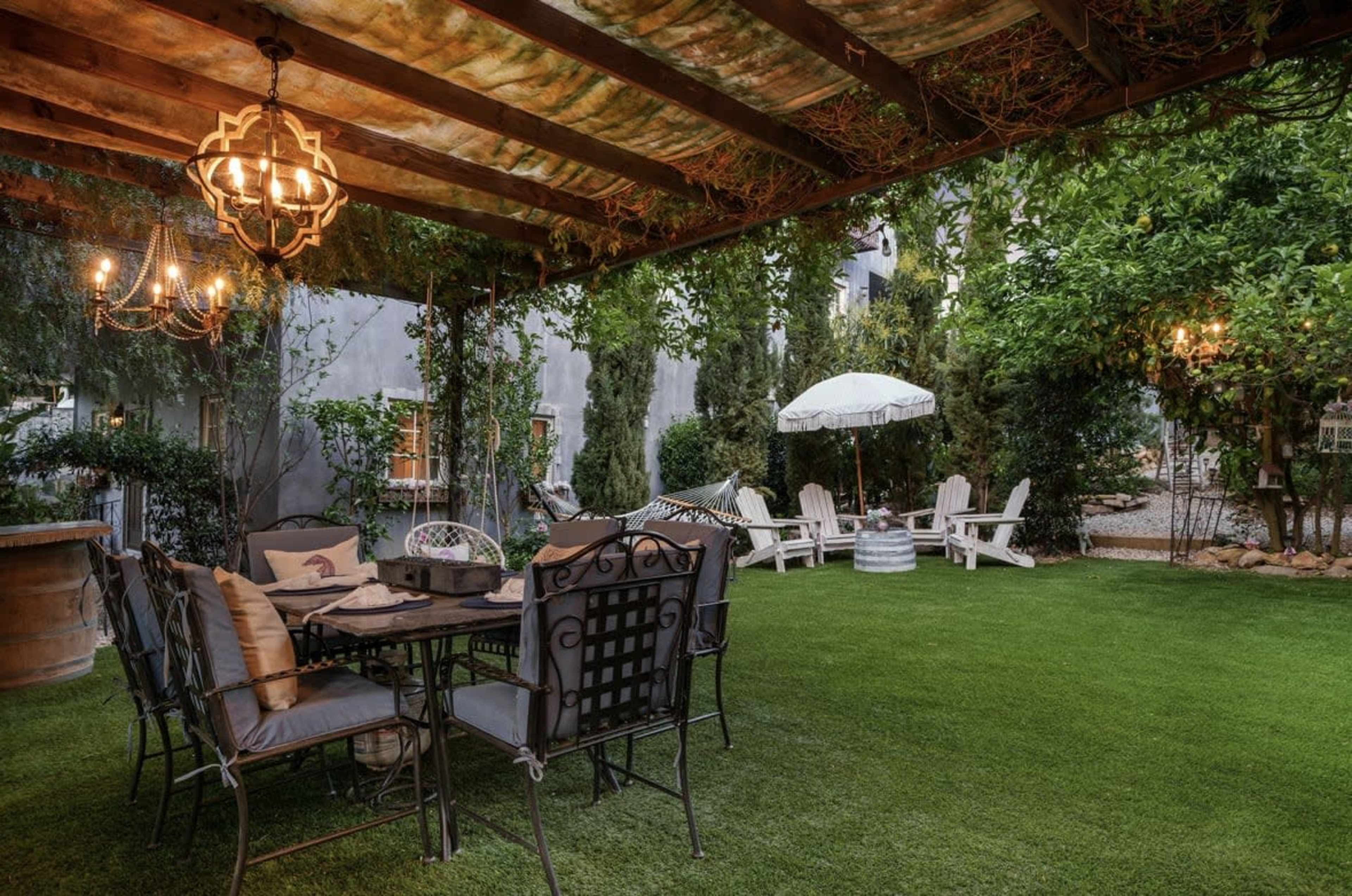 The image shows a cozy outdoor space with a dining table and chairs under a wooden pergola, surrounded by lush greenery and a hammock.