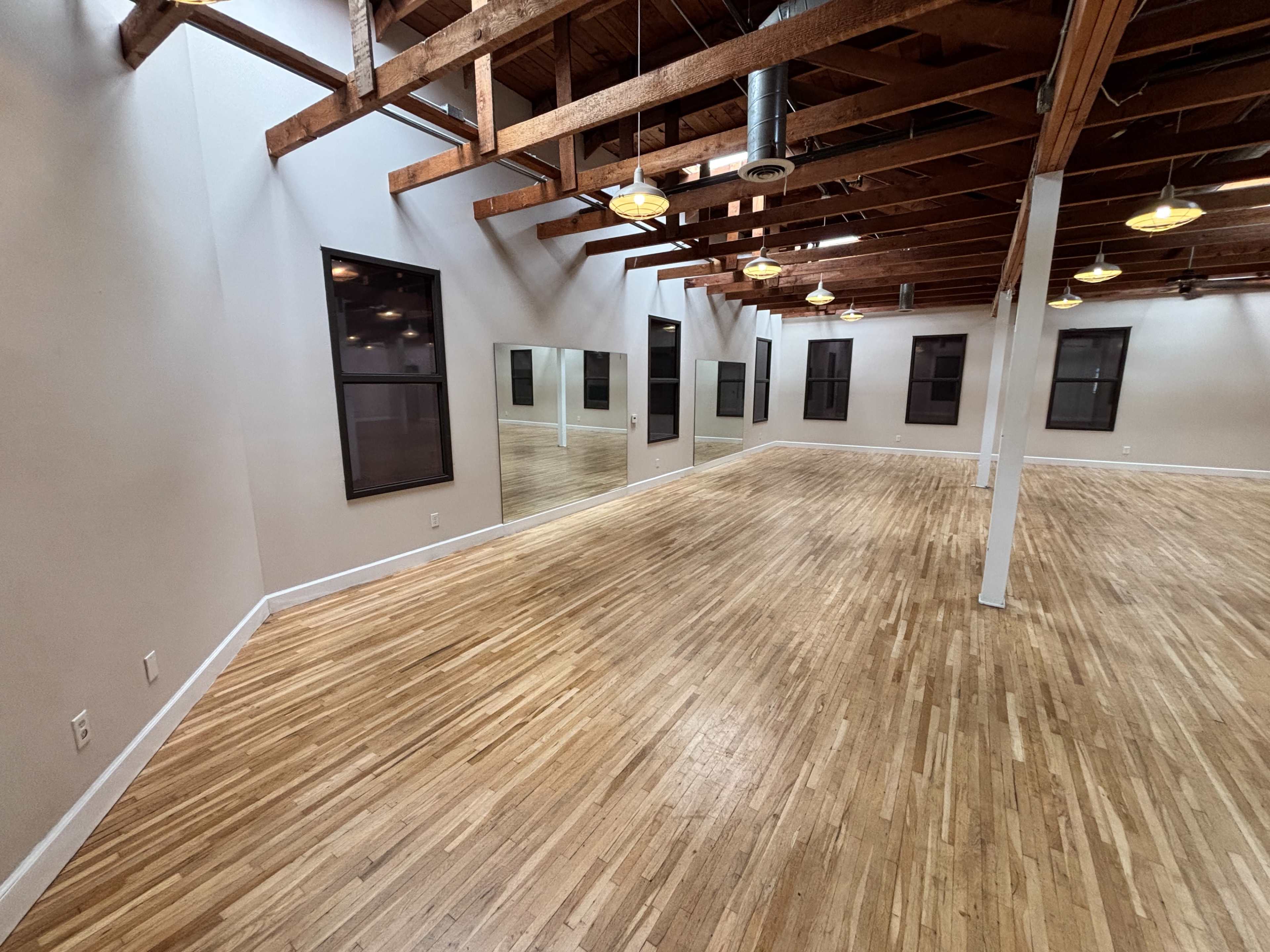 The image shows an empty studio space with hardwood floors, exposed wooden beams, and large windows, featuring a mirror along one wall.