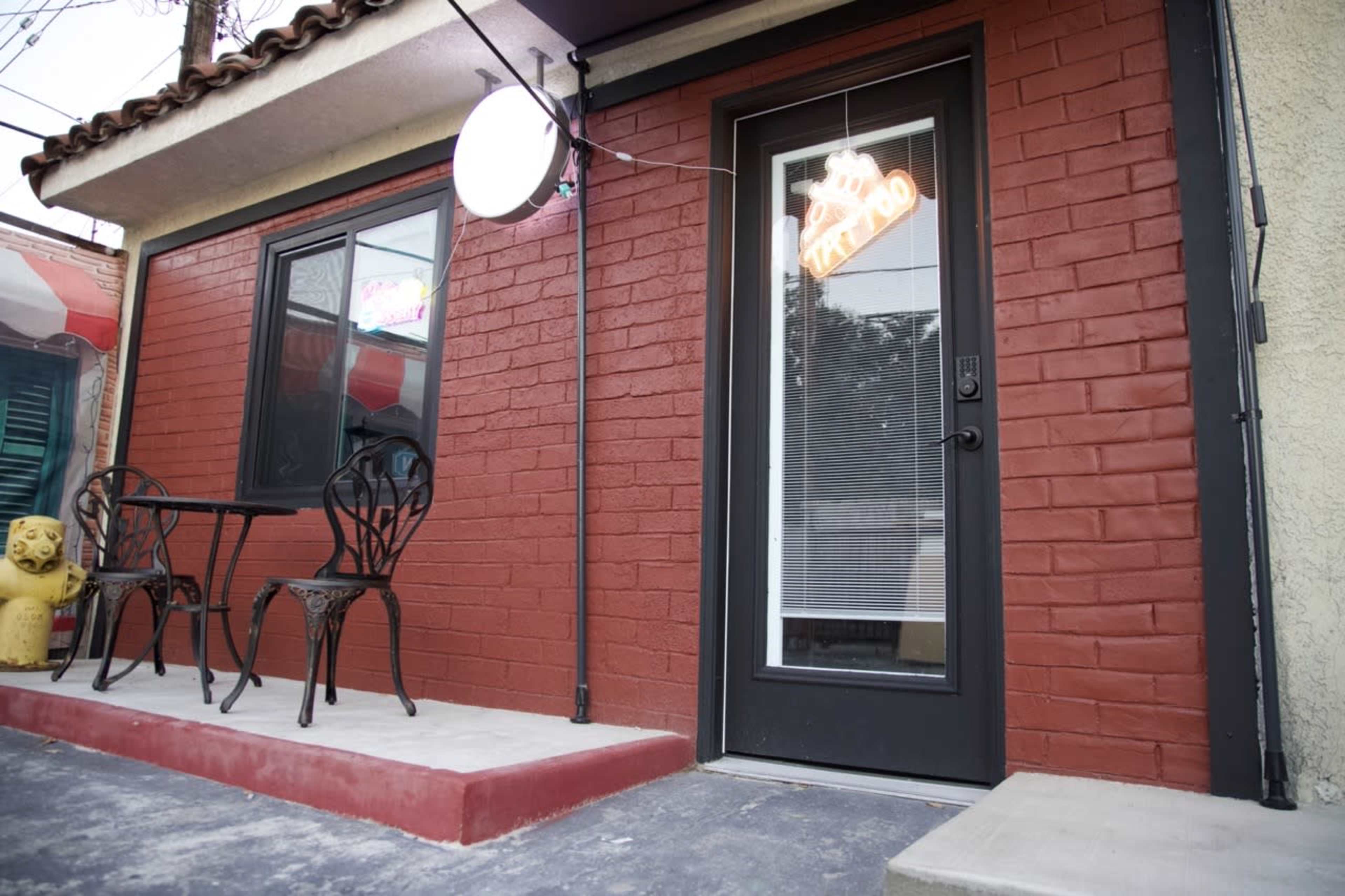 The image shows a brick building with a storefront featuring a "Tattoo" neon sign, a small table with two chairs, and a decorative lion statue nearby.