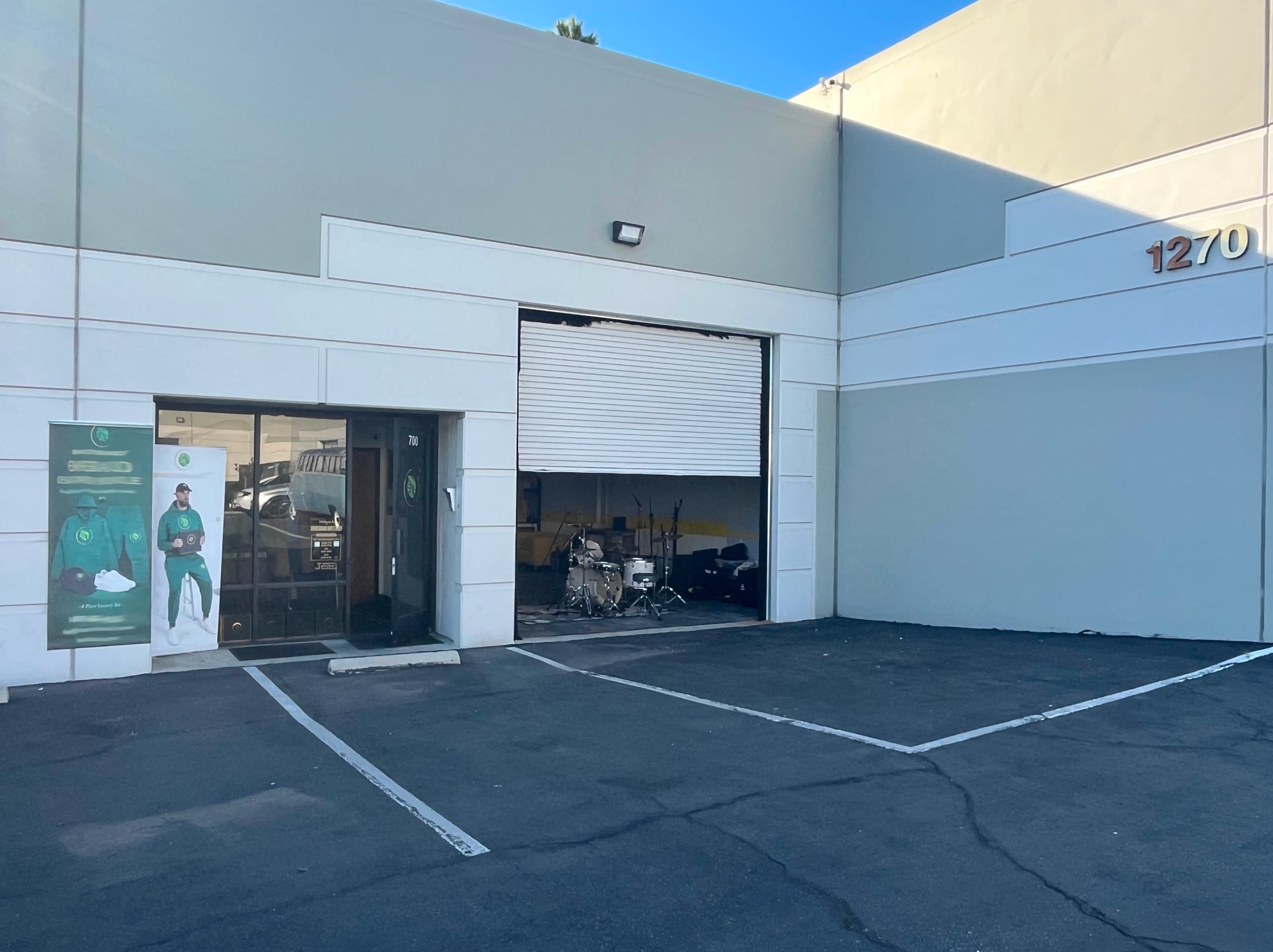 The image shows a commercial building with a large open garage door revealing a drum set inside, along with a glass entrance to the left.