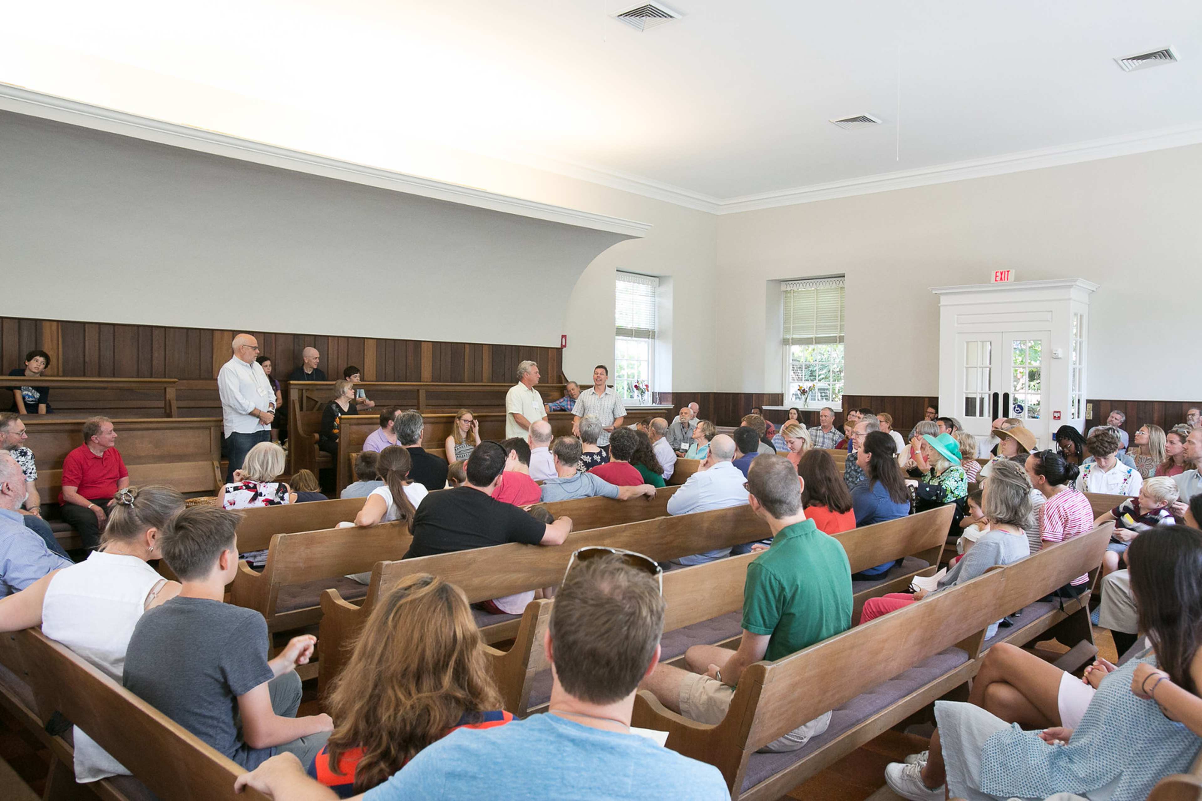 A crowd of people is seated in a wooden meeting hall while speakers stand at the front.