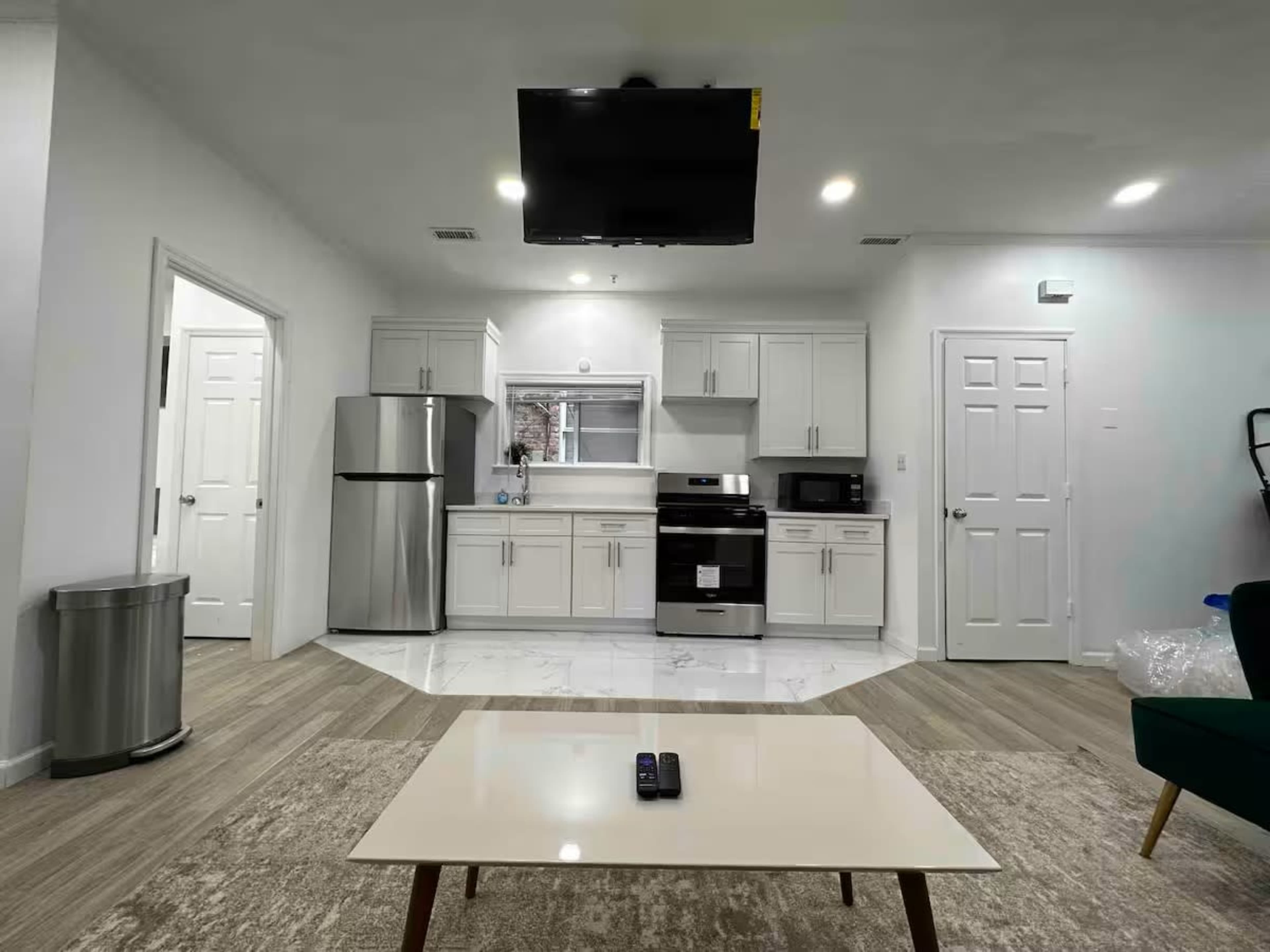 The image shows a modern kitchen and living area with a stainless steel refrigerator, stove, and a television mounted on the wall.