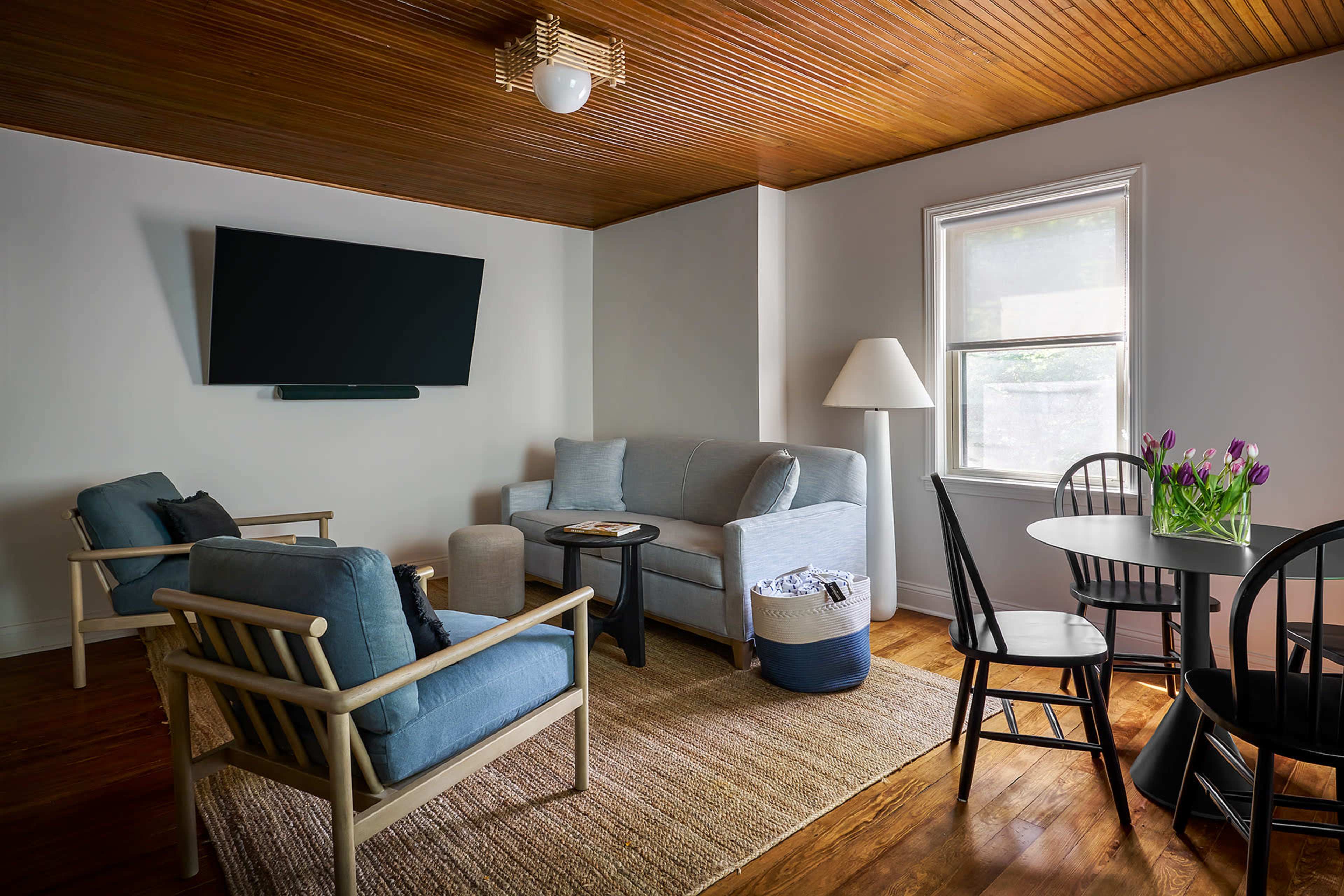 A cozy living room features a gray sofa, two armchairs, a dining table, and a window, all set against a wooden ceiling and warm hardwood floors.