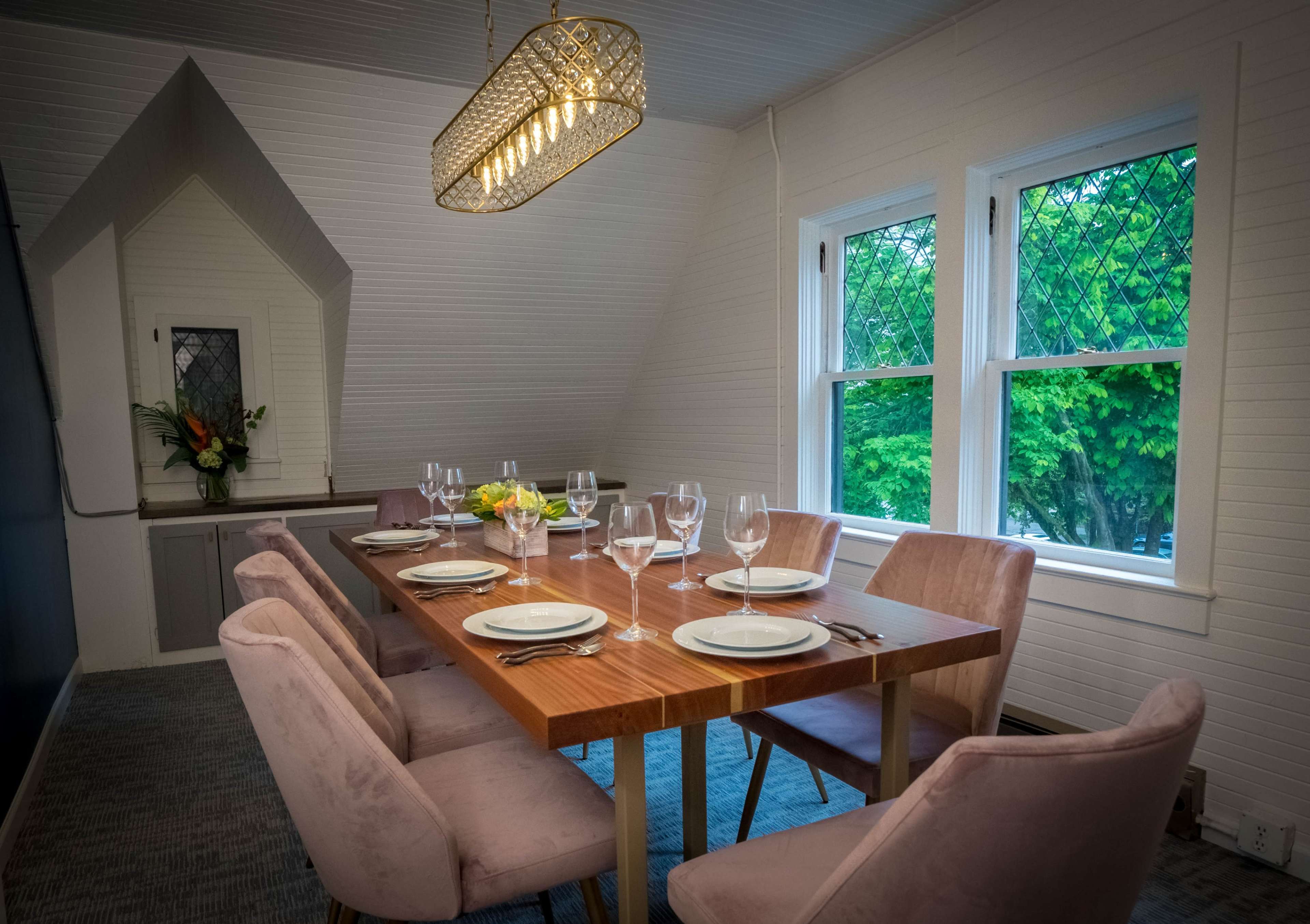 A modern dining room features a long wooden table set for six with pink chairs and large windows allowing natural light and views of greenery outside.