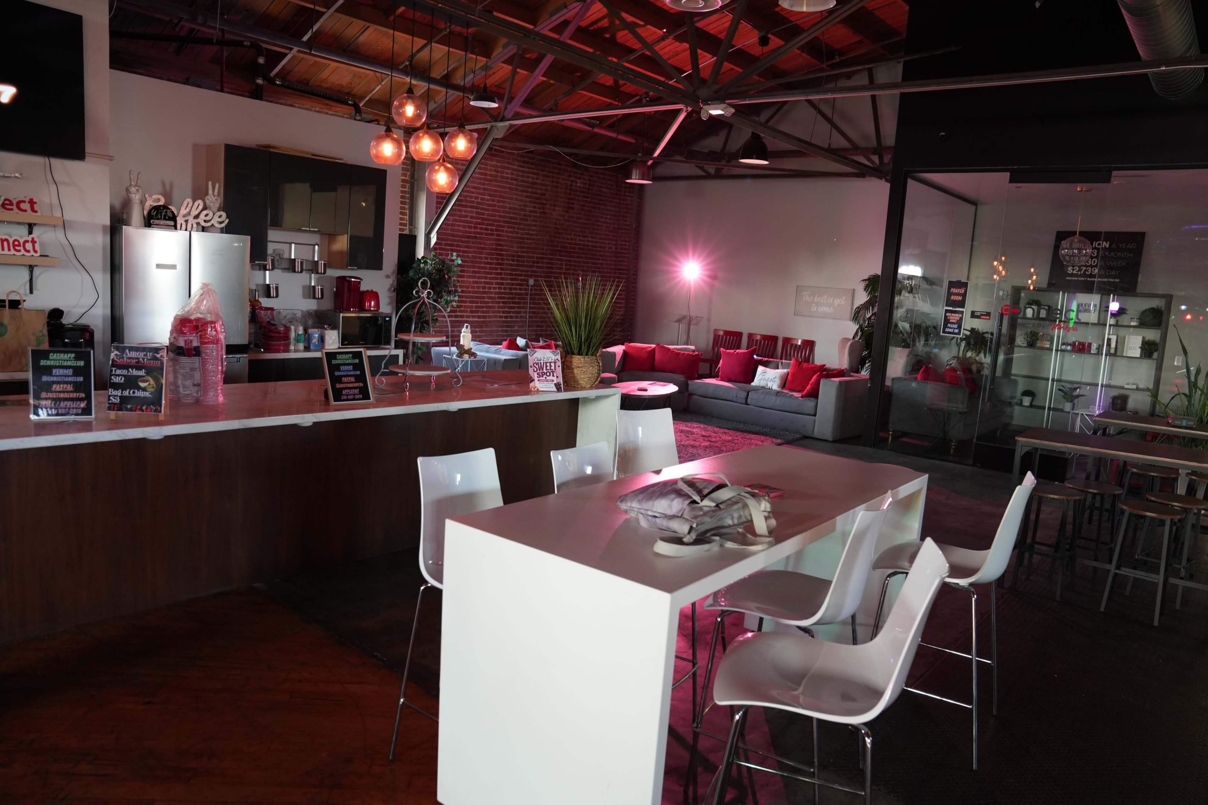 The image shows a modern, open-concept café area with white furniture, a bar counter, and a lounge space featuring a plush sofa and decorative plants.