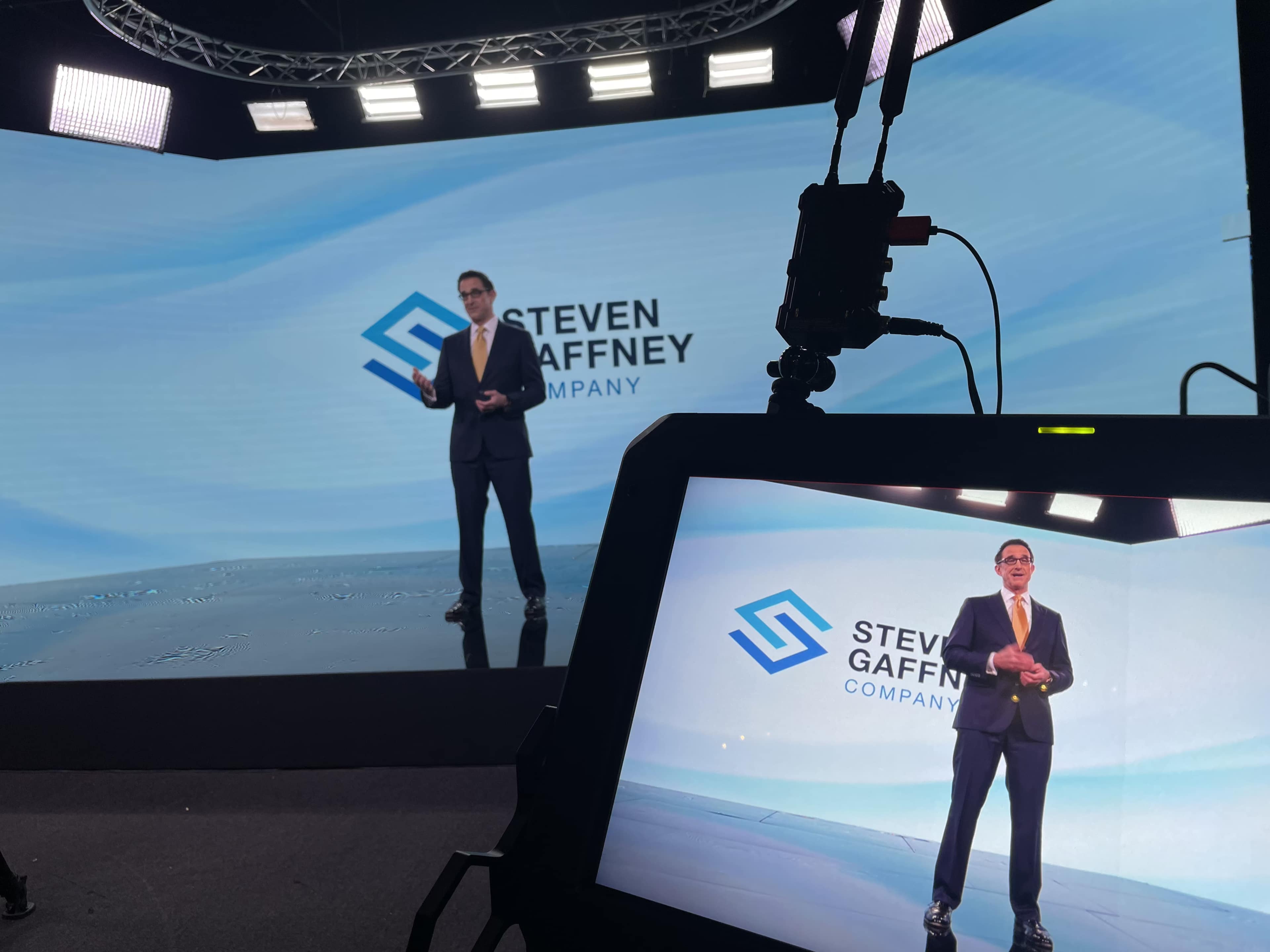 A presenter stands on a stage with two large screens displaying the name "Steven Gaffney Company" in the background.