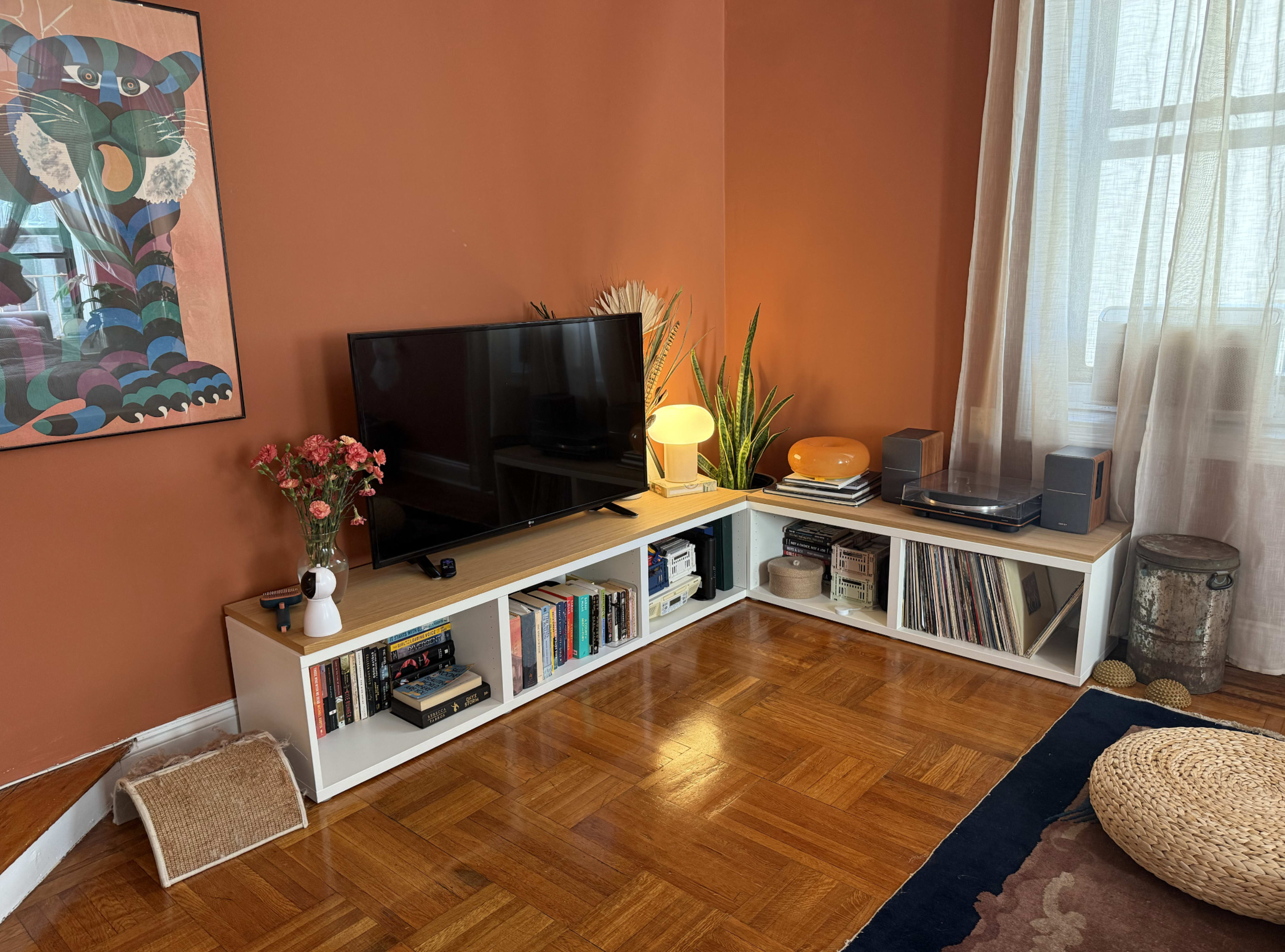 The image shows a living room with an orange wall, a TV on a white media console, bookshelves filled with books, a lamp, a turntable, and plants.