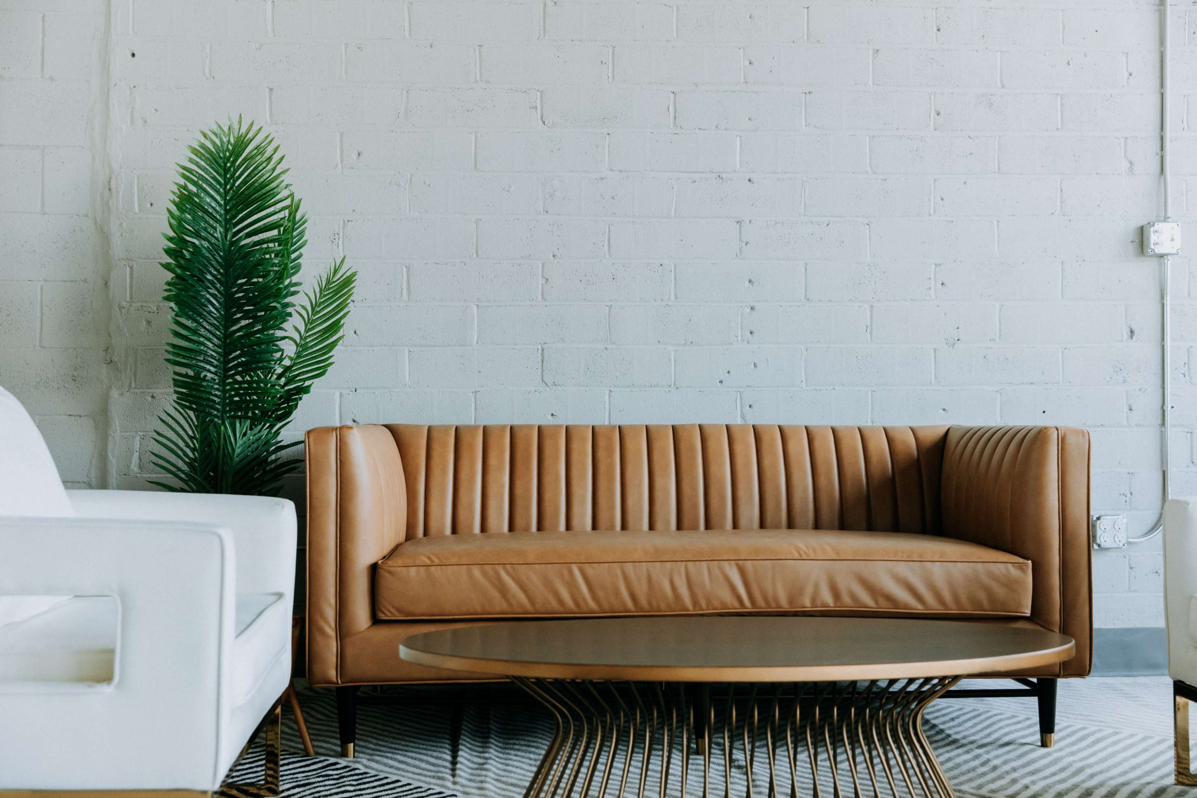 A brown leather couch with vertical ribbing sits in front of a white brick wall, accompanied by a round coffee table and a green potted plant.