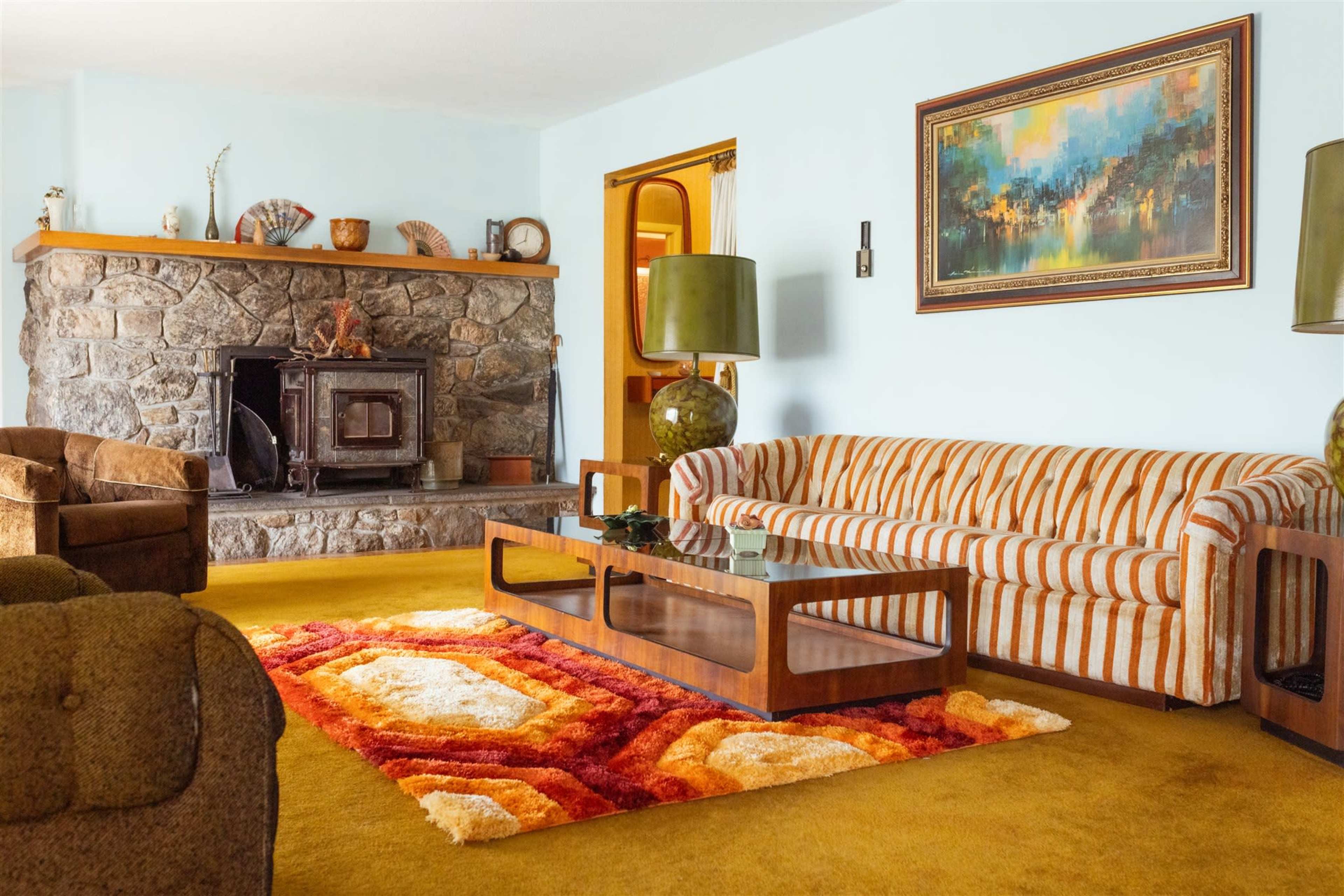 The living room features a stone fireplace, a striped sofa, and a colorful area rug with a glass coffee table in the center.