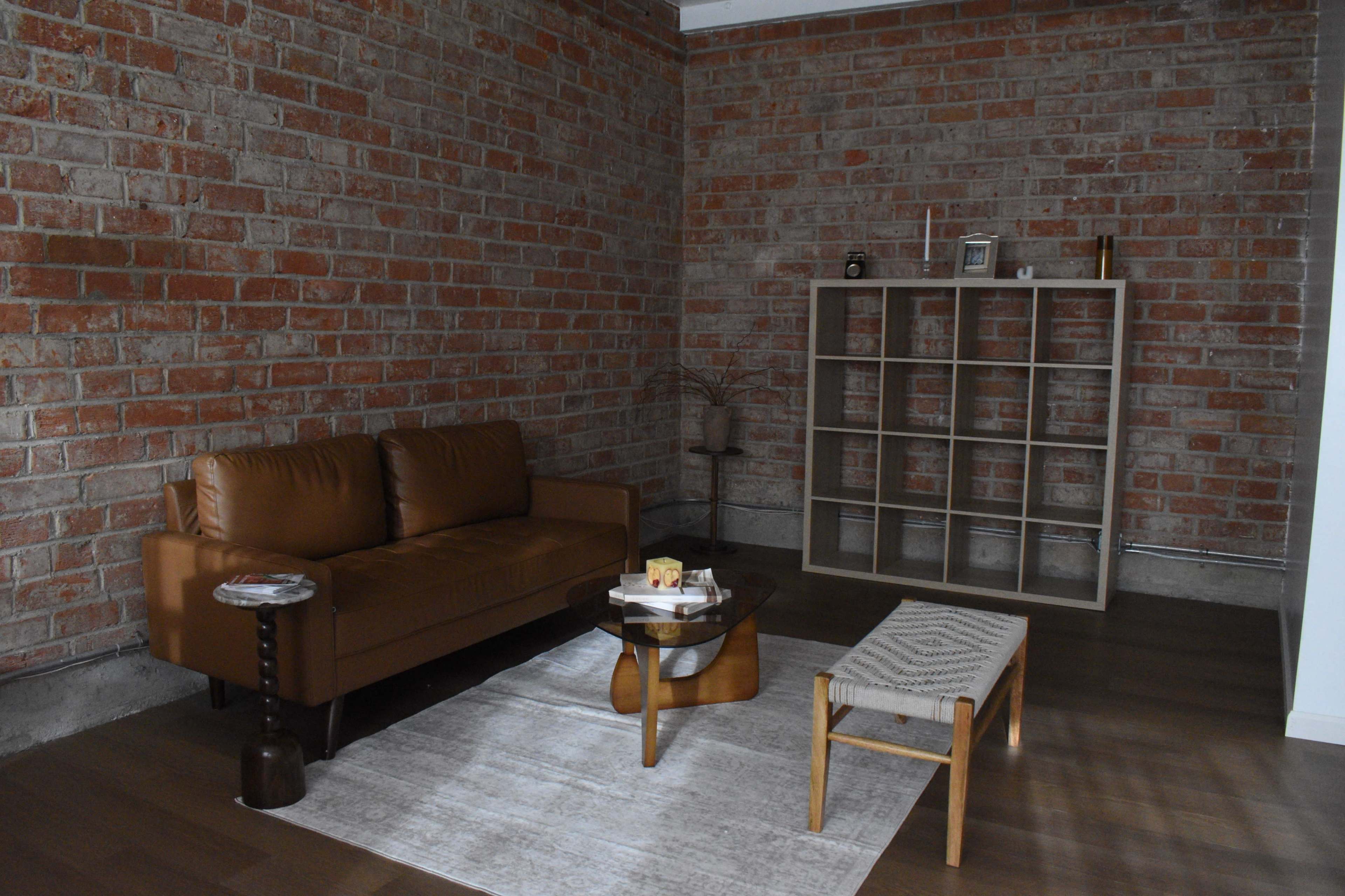 The room features a leather sofa, a coffee table, a woven bench, and an open shelving unit against a brick wall.