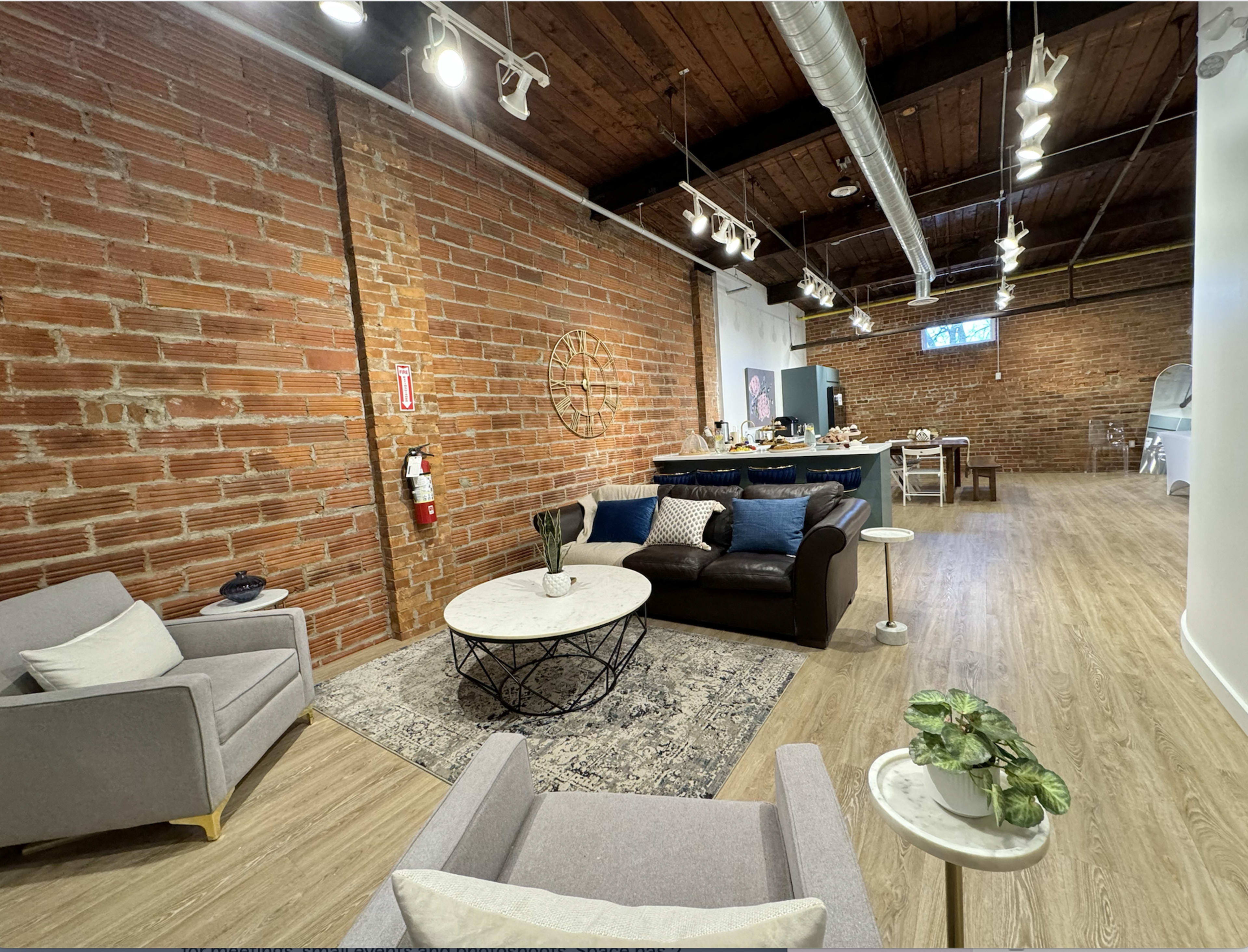 The image shows a spacious living area with exposed brick walls, a modern sofa, a coffee table, and a dining area in the background.