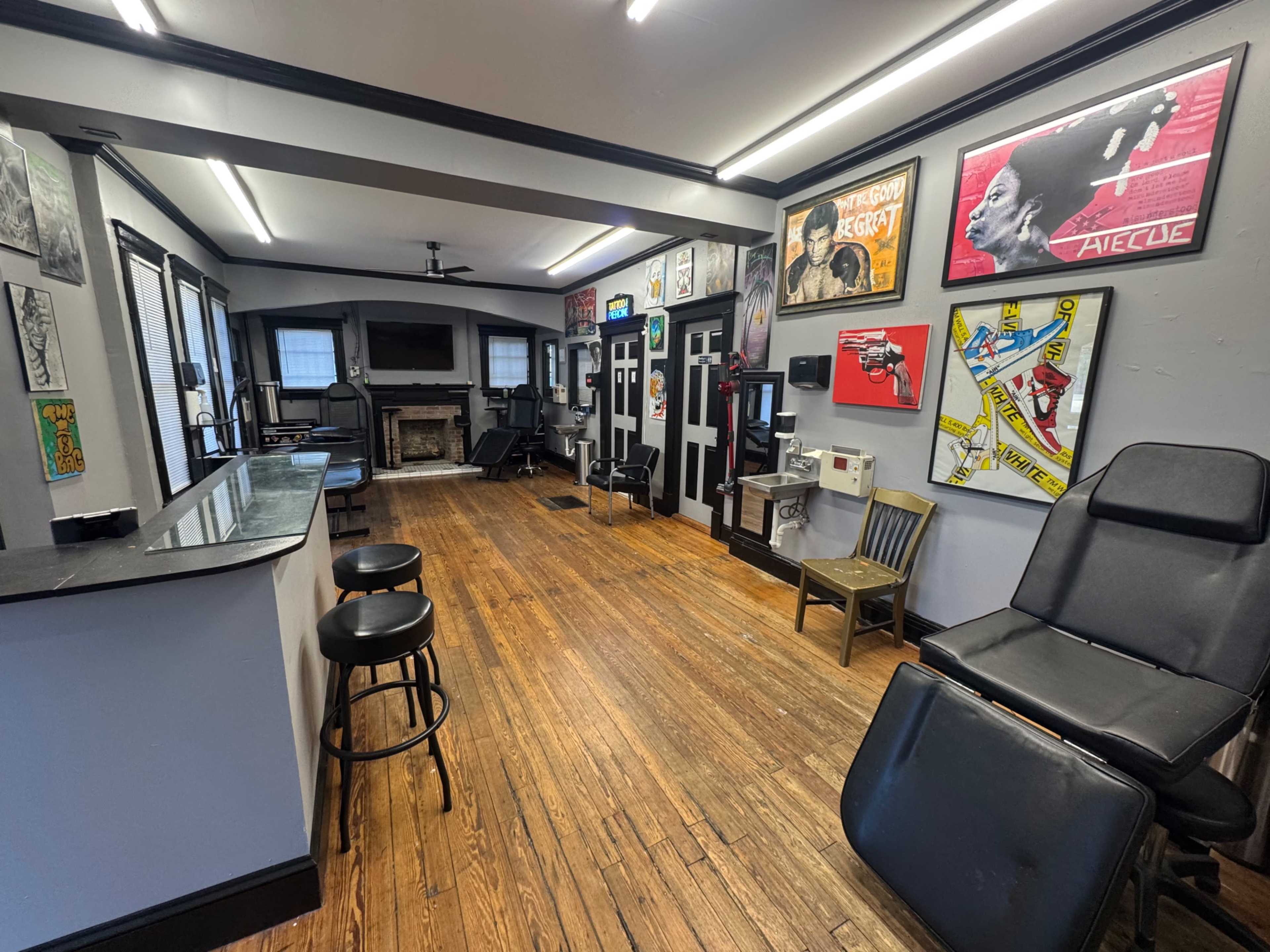 The image shows an interior of a barber shop with wooden floors, a reception area, and walls adorned with framed artwork and posters.