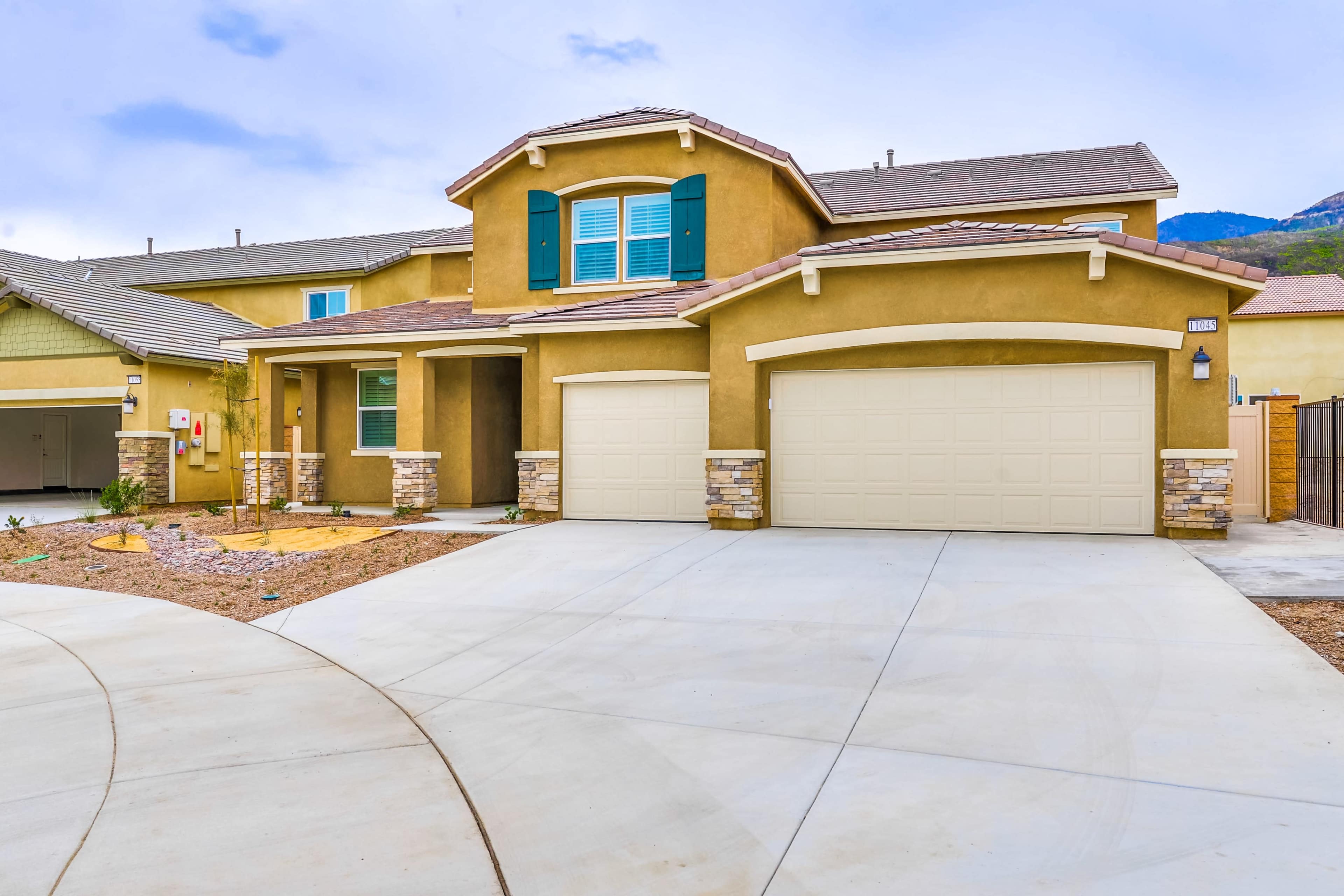 A two-story house with a beige exterior, green shutters, and a double garage, situated on a paved driveway.
